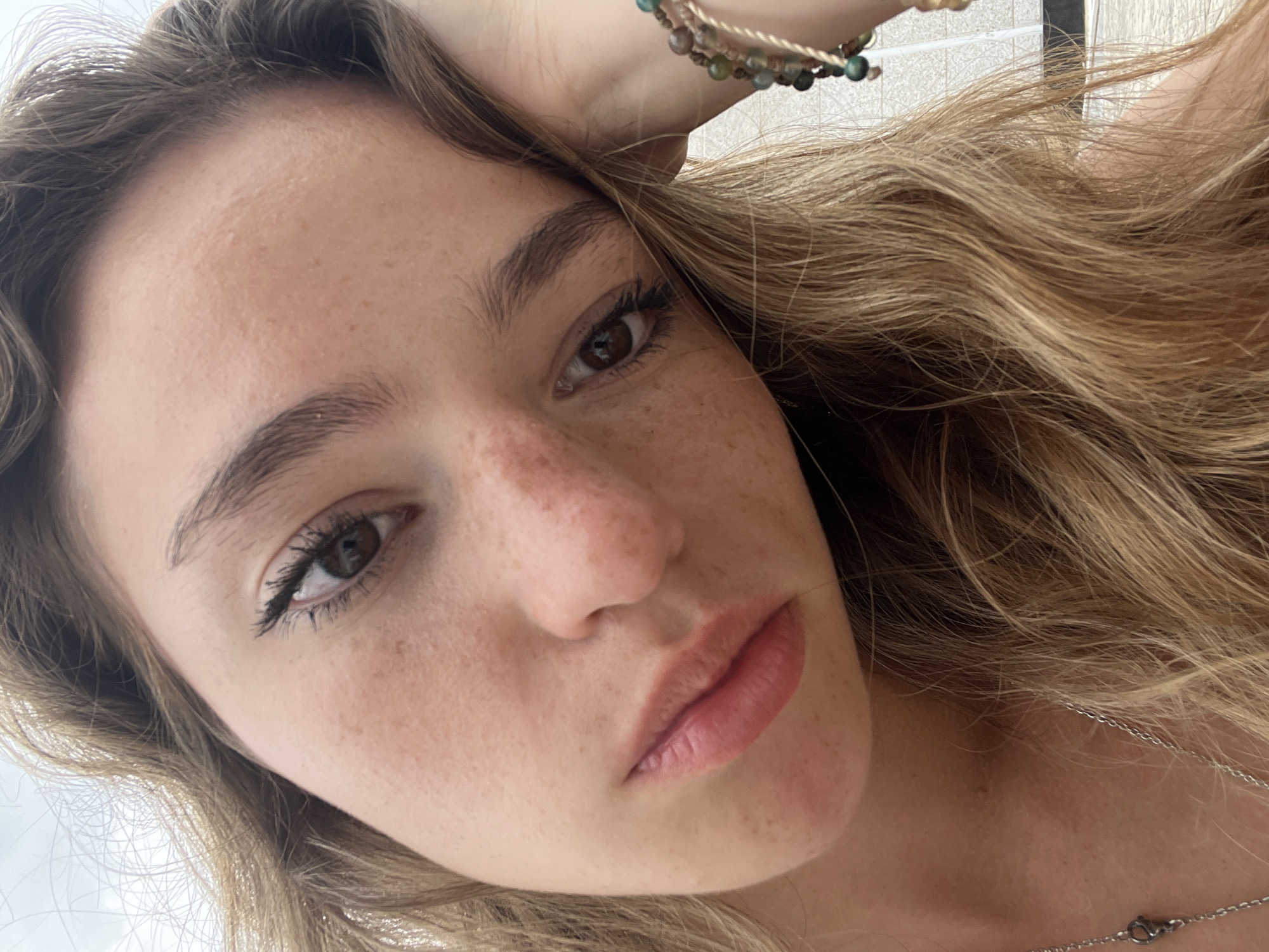 A young woman with fair skin, freckles, and light brown hair is facing the camera. Her hair is partially covering her forehead and parts of her face. She has brown eyes and full lips. A beaded bracelet is visible on her left wrist. A silver necklace is also visible around her neck.
The background is indistinct and does not provide specific information to identify the location as Puebla, Mexico.