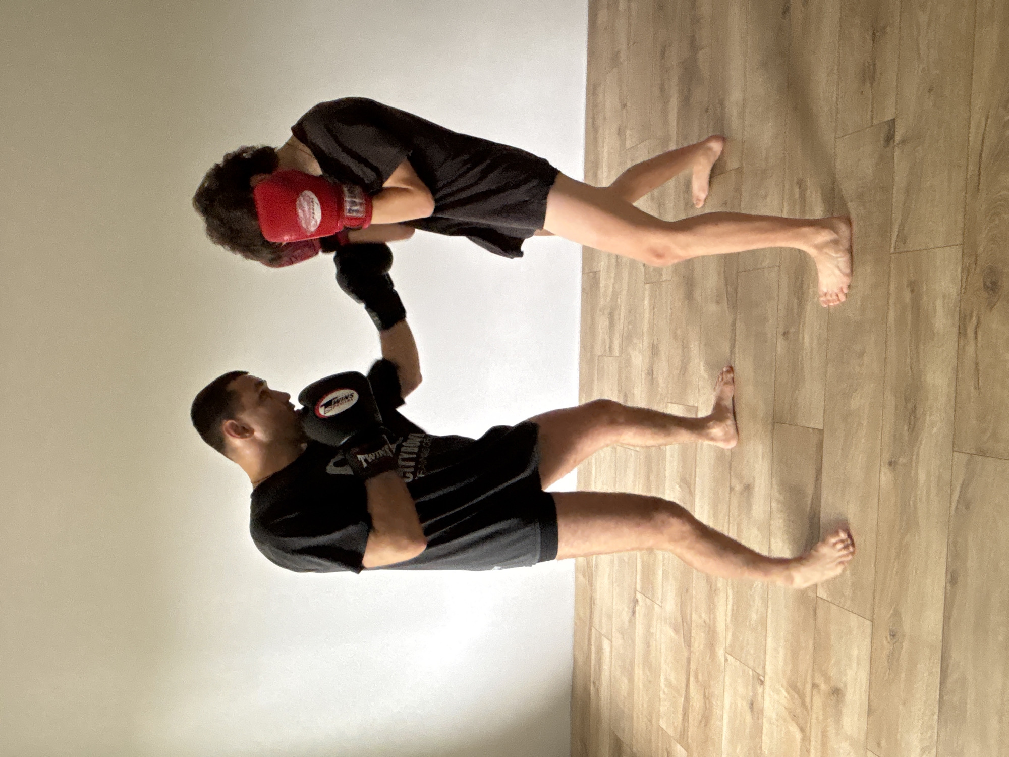 The image depicts two individuals engaged in a combat sports training session indoors, likely in Chaikovsky, Russia. The scene, when correctly oriented, shows two men dressed in black t-shirts and black shorts, both barefoot, actively sparring.
On the left, one man wears black boxing gloves, featuring the "TWINS" brand logo prominently on the back and "TWINS FIGHTGEAR" on the wrist strap. He is captured in the motion of delivering an upward strike, with his right arm bent and glove near the other person's head.
On the right, the second man, with dark, curly hair, wears red boxing gloves, also bearing the "TWINS" logo. He is in a defensive stance, with both red-gloved hands raised to protect his face and head, indicating he is either blocking or bracing for a strike.
The setting is a simple indoor room with plain, light-colored walls and a light brown, wood-effect laminate floor. The lighting is even, suggesting either artificial light or indirect natural light. The focus is entirely on the training activity, with no other objects or furniture visible, lending an uncluttered and functional atmosphere to the training space. The time of day appears to be daytime.