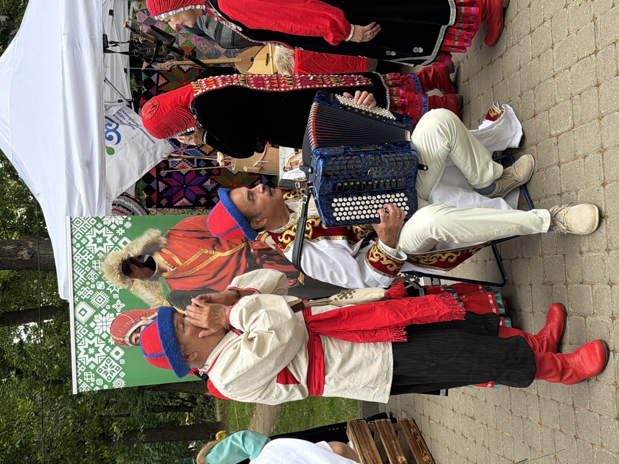 The image captures a group of people in traditional attire, likely at an outdoor cultural event or festival in Kazan, Russia. The scene is dominated by vibrant colors and intricate patterns.
In the foreground, a man wearing a red and blue hat, a white tunic with red and gold embroidery, and black pants with a red sash is playing a blue accordion. He is seated, and his eyes are closed as if deeply engrossed in the music. Next to him, another person, also in traditional clothing, is partially visible, with their hands clasped.
Behind them, a woman in a red headdress and a black garment with ornate red and white trim is also present. Further back, a large banner or backdrop features a stylized green pattern and an image of someone in traditional attire with a fur-lined collar. The setting appears to be outdoors, with cobblestone ground visible and trees in the background, suggesting a public space like a park or a market square.
The attire suggests a cultural performance or demonstration, possibly related to Tatar or other regional traditions. The atmosphere seems festive, with a focus on music and traditional craftsmanship. The time of day appears to be daytime, and the weather seems pleasant. The overall impression is one of cultural celebration and artistic expression. No discernible text is clearly readable in the image, although some writing is visible on a white banner.