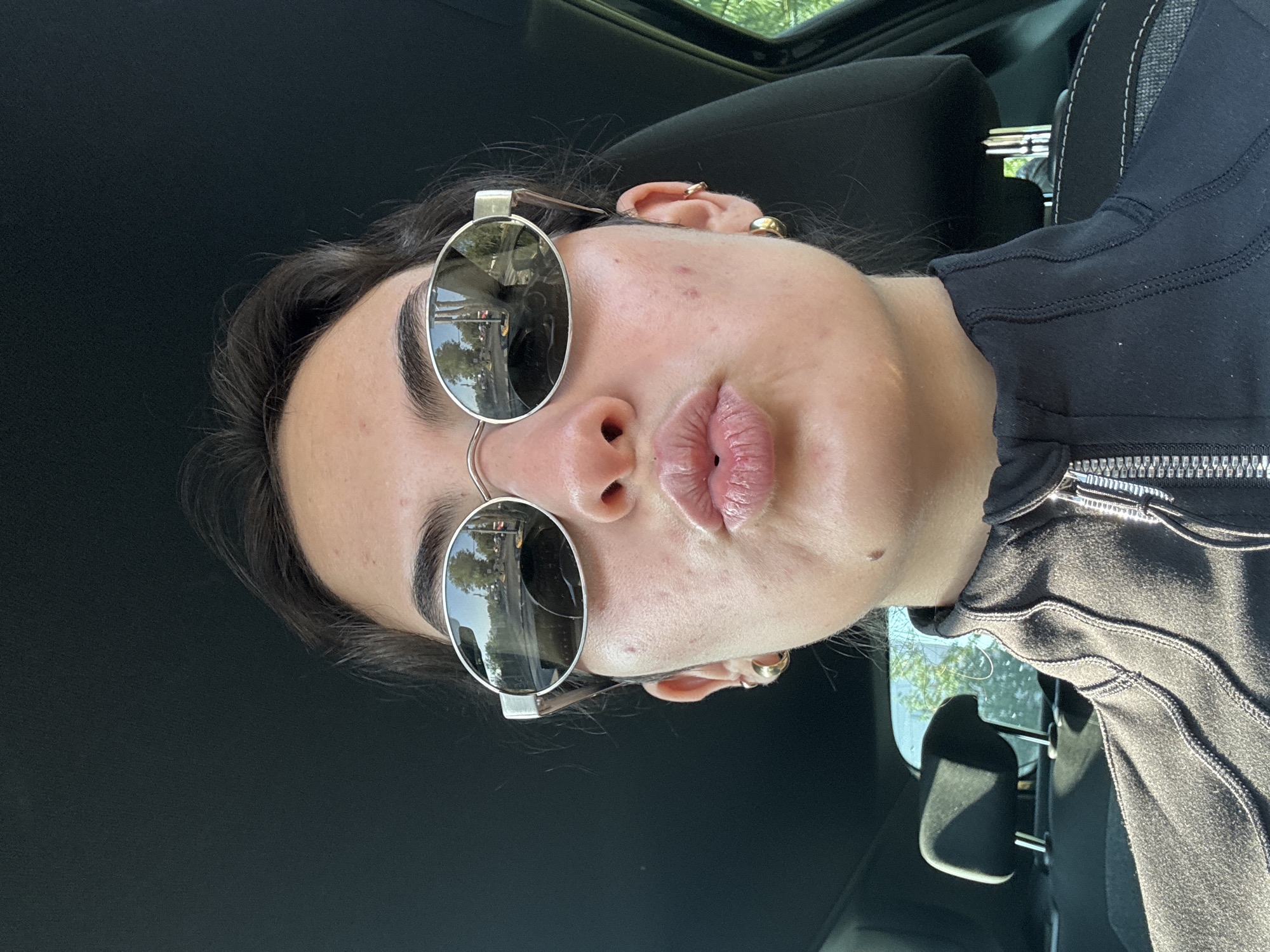 A close-up selfie shows a person with light skin and dark hair. The person is wearing round, silver-framed sunglasses that reflect a street scene. The location appears to be Puebla, Mexico based on the architecture visible in the reflection of the sunglasses. The person is also wearing gold earrings. The background is primarily black, suggesting the interior of a vehicle. The person's lips are slightly puckered. The person is wearing a black zippered top. The scene is well-lit with natural sunlight.