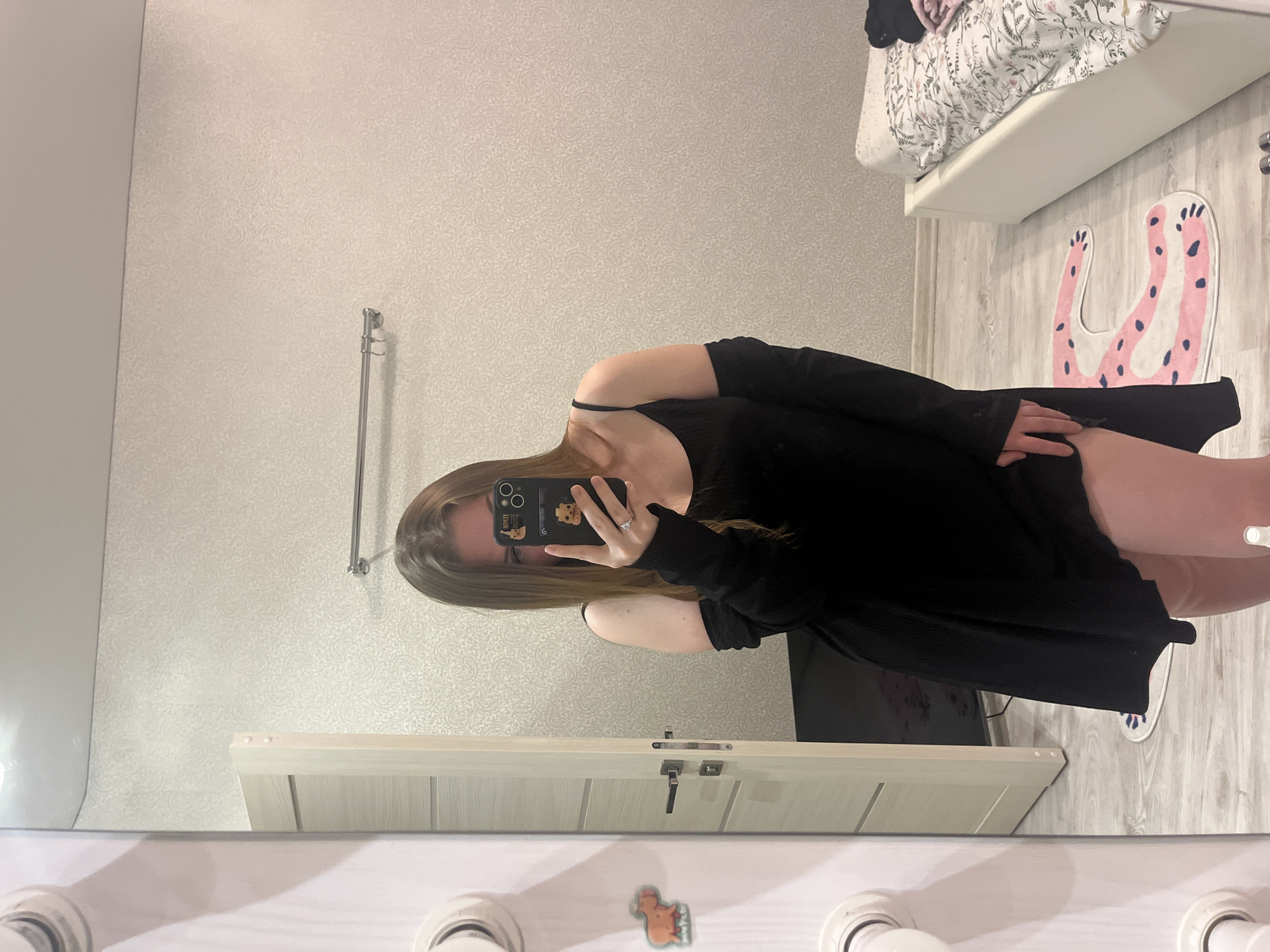 A woman stands in front of a bathroom mirror taking a selfie. She wears a black, long-sleeved top. The room includes a towel rack, a bed, a rug shaped like animal paws, a dresser, and a television. The location is not discernible as Sevastopol, Ukraine, from the image.