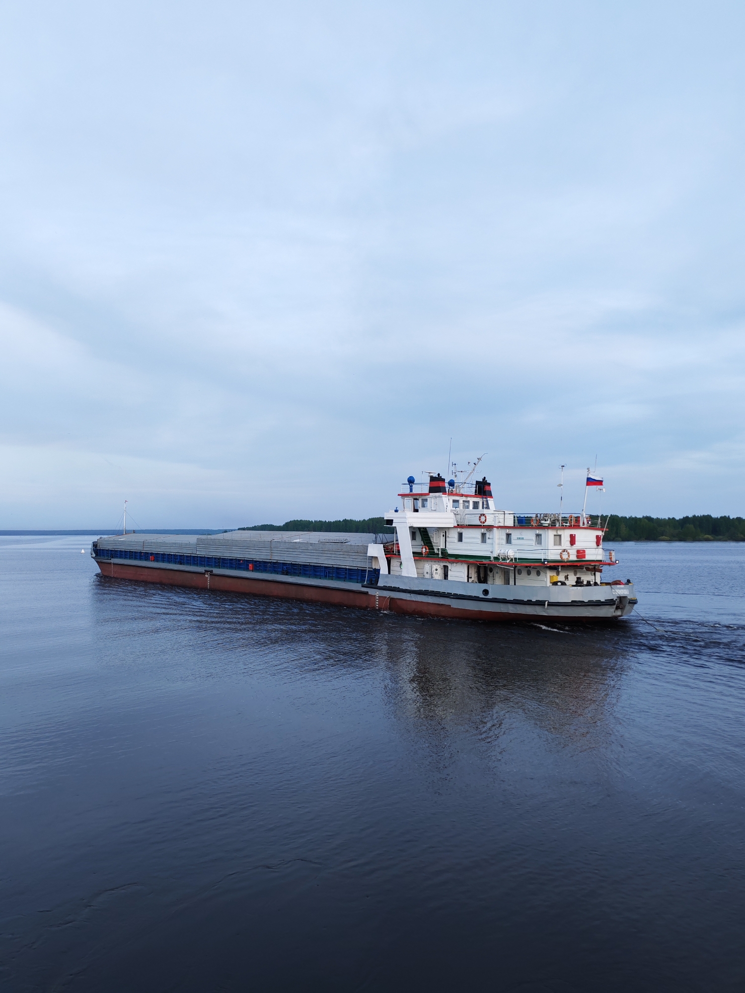 A barge travels on a body of water under an overcast sky. The barge is composed of two parts: a flat-topped, rectangular cargo carrier, and a smaller, multi-tiered tugboat. The cargo carrier has a grey top, blue sides, and a red-brown bottom. The tugboat, attached to the carrier’s rear, is white with grey accents, featuring red railings and details. A Russian flag flies from the tugboat. The water is dark and reflective, showing the barge's reflection, with a wake trailing the vessel. A wooded shoreline is visible in the distance on the right side of the image.