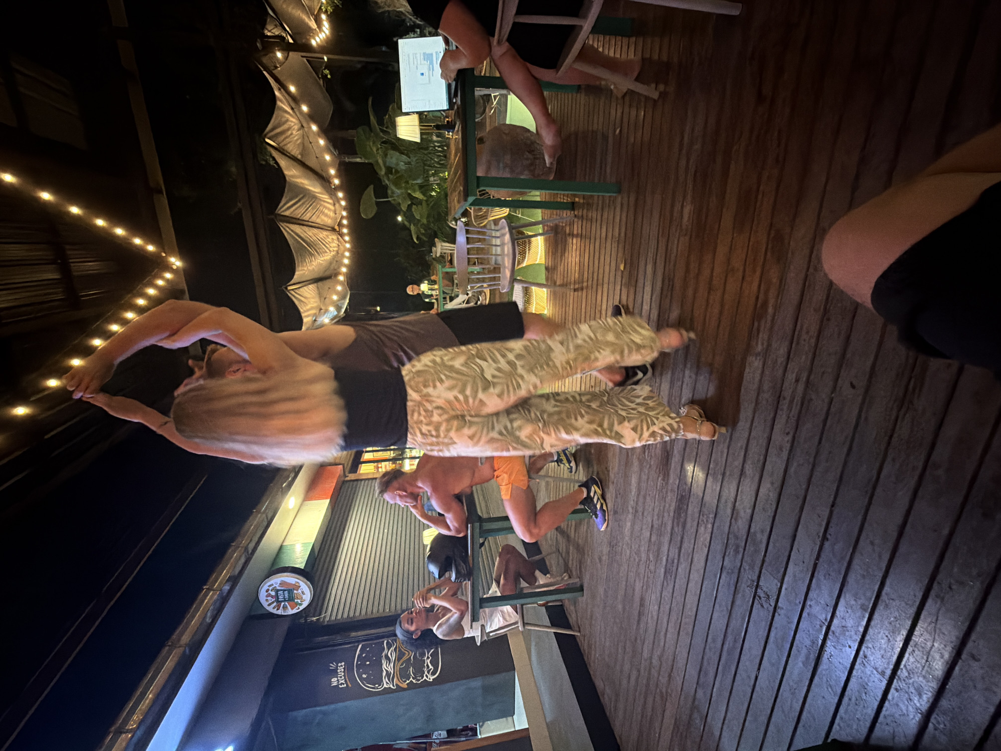 A night scene shows a couple dancing in the foreground of a restaurant in Ban Rak, Thailand. The woman, with long blonde hair, wears a black top and patterned pants. The man, behind her, is wearing a dark tank top and shorts. In the background, two people are seated at a table. A woman in a white dress sits on the left, and a shirtless man in orange shorts is on the right. A motorcycle helmet rests on their table. String lights hang from the ceiling above the outdoor seating area, illuminating the wooden deck. Further back, a woman is seated at a table with a laptop, and tropical plants are visible. The restaurant has a dark facade with a sign featuring a hamburger logo and the words "NO EXCUSES." The overall ambiance is casual and lively.