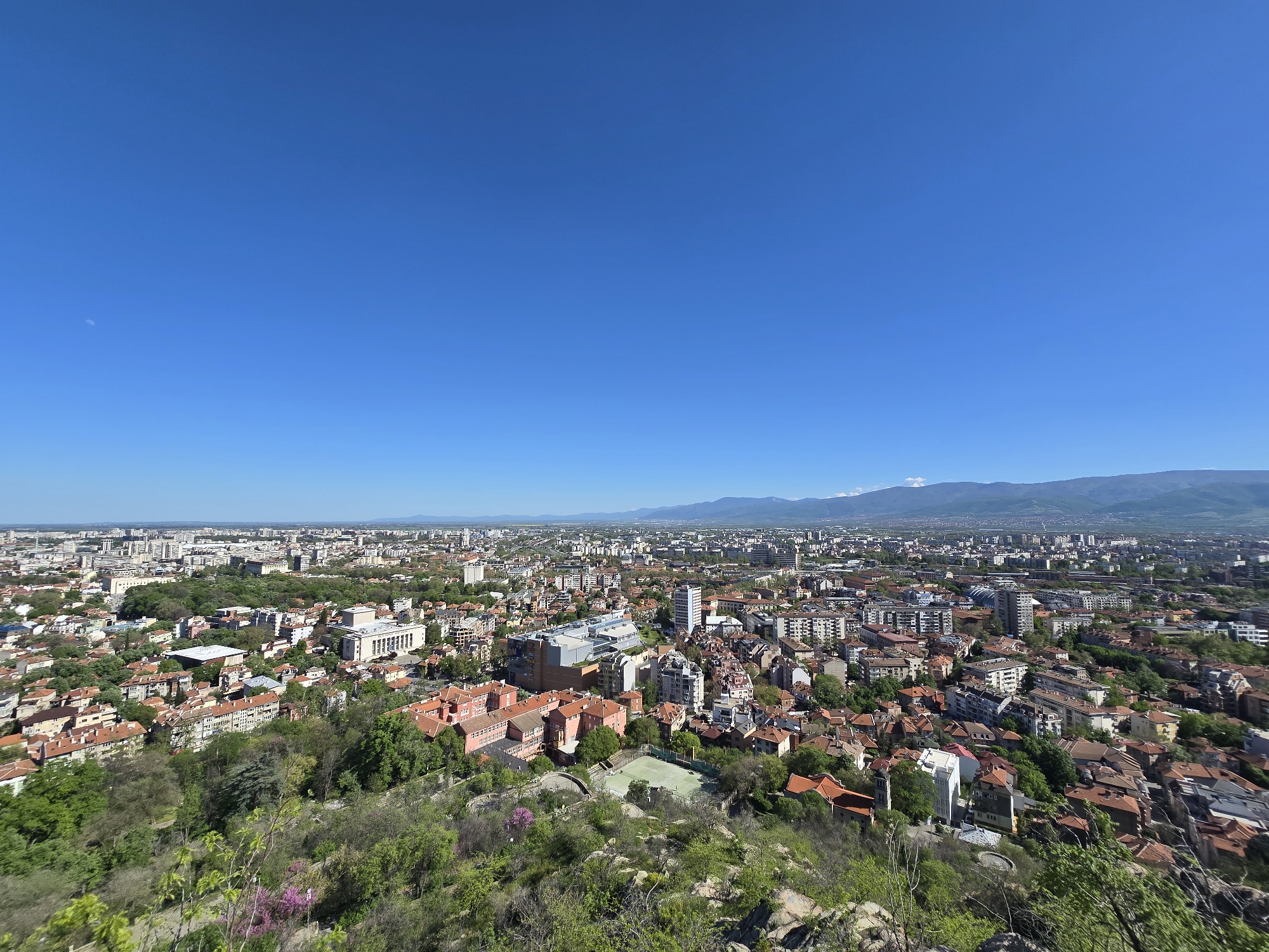 The image presents a wide-angle aerial view of a city under a clear blue sky. In the foreground, there is a rocky outcrop with greenery. The city spans across the middle ground, with a mix of residential and commercial buildings. Red-tiled roofs are prevalent in the lower sections, transitioning to taller, modern structures towards the horizon. Mountains are visible in the distant background.
