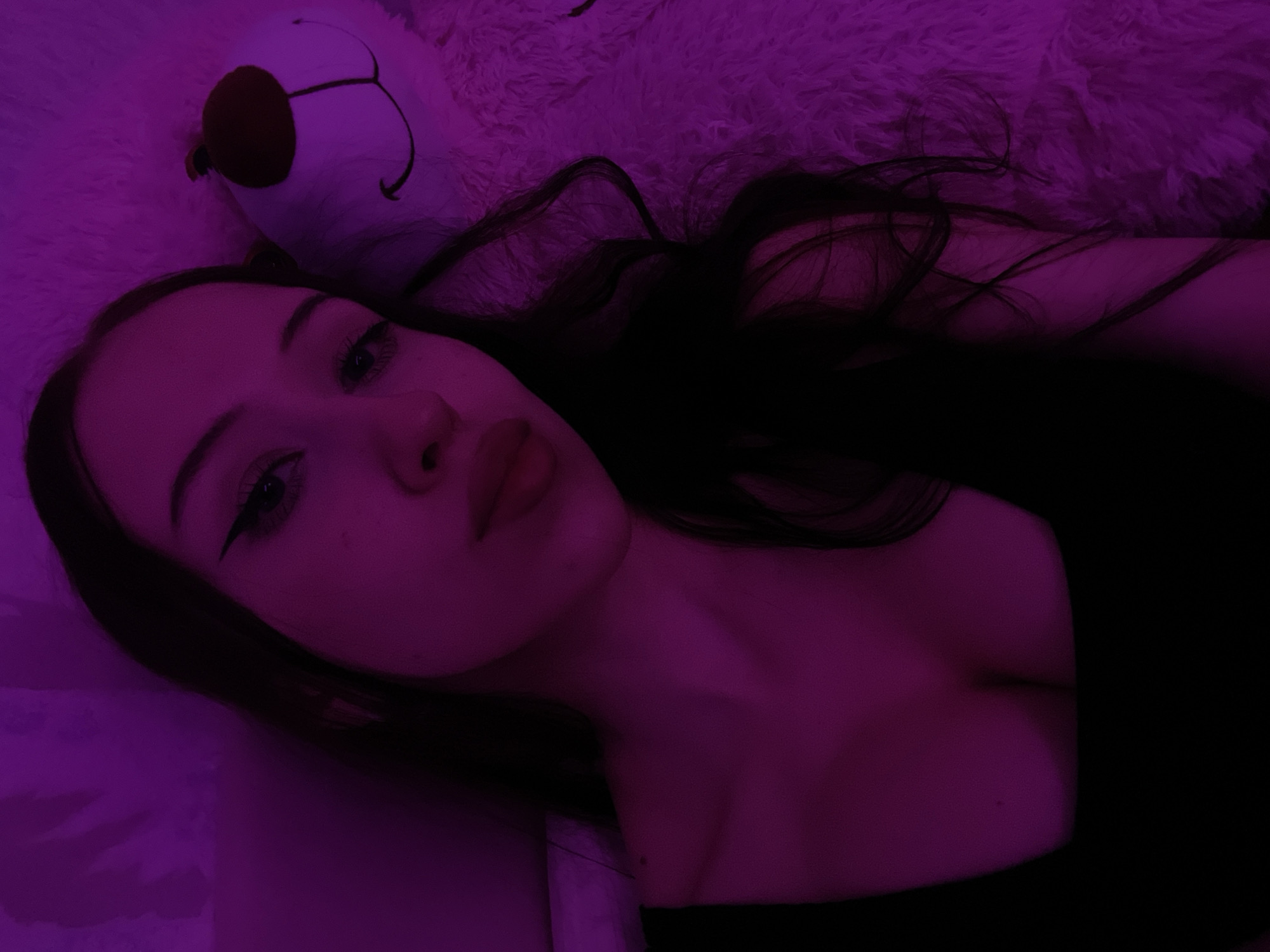 The image is a close-up, low-angle selfie of a young woman with long, dark hair and winged eyeliner. She is wearing a black top that reveals her décolletage. To her right is a fluffy, white teddy bear with a red nose and a black smile. The scene is illuminated by a vibrant purple light, creating a moody atmosphere. The woman is lying down, as indicated by the soft, textured surface she is resting on, which is likely a blanket or rug. The background is indistinct due to the lighting and focus. No specific location in Votkinsk, Russia, can be identified from the image.