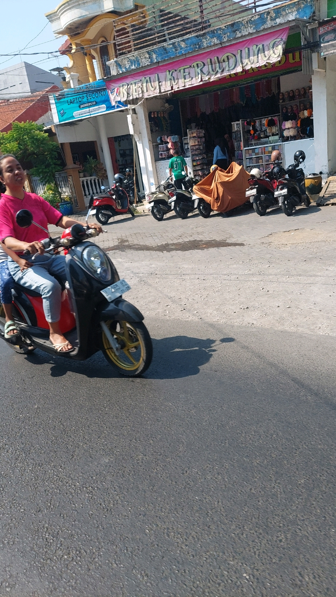 The image depicts a street scene in Sidoarjo, Indonesia. In the foreground, a woman wearing a pink shirt and blue jeans is riding a black scooter with a red accent. She is accompanied by a passenger. The scooter has a license plate that reads "W 3355 D Z". Behind them, several other scooters are parked on the side of the road. A store with a banner that reads "RUMAH KERUDUNG" is visible, selling various items, possibly clothing or accessories. The architecture suggests a commercial area in a city or town. The overall environment is sunny, with clear skies.