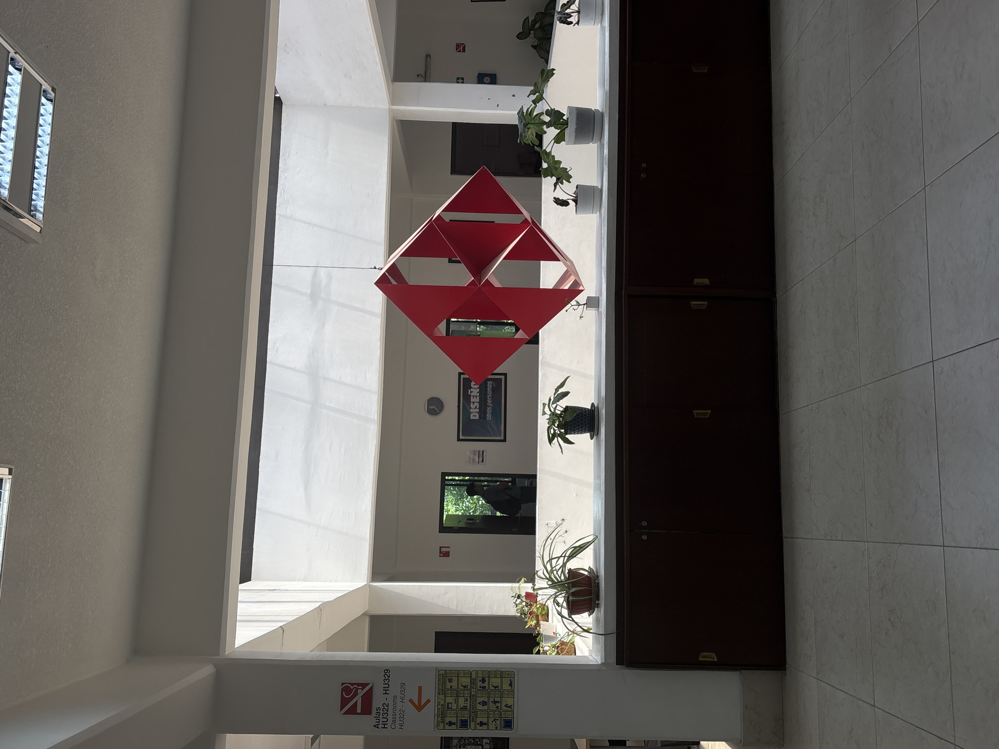 The image shows an interior space in San Andrés Cholula, Mexico, featuring a red, geometric, three-dimensional sculpture suspended from the ceiling in the foreground. The space appears to be a hallway or atrium. Natural light streams in through windows, illuminating white walls and structural supports. Potted plants line a ledge along the wall on the right side of the image, beside dark wooden cabinets. A sign is visible on a pillar near the bottom of the frame with the words "Aulas" and "HU322 - HU329" alongside pictograms, indicating classrooms. Further down the hallway, signs and a framed poster with the word "DISEÑO" (design) are seen. In the background, a person is visible within an open doorway. The floor is tiled in a neutral color.