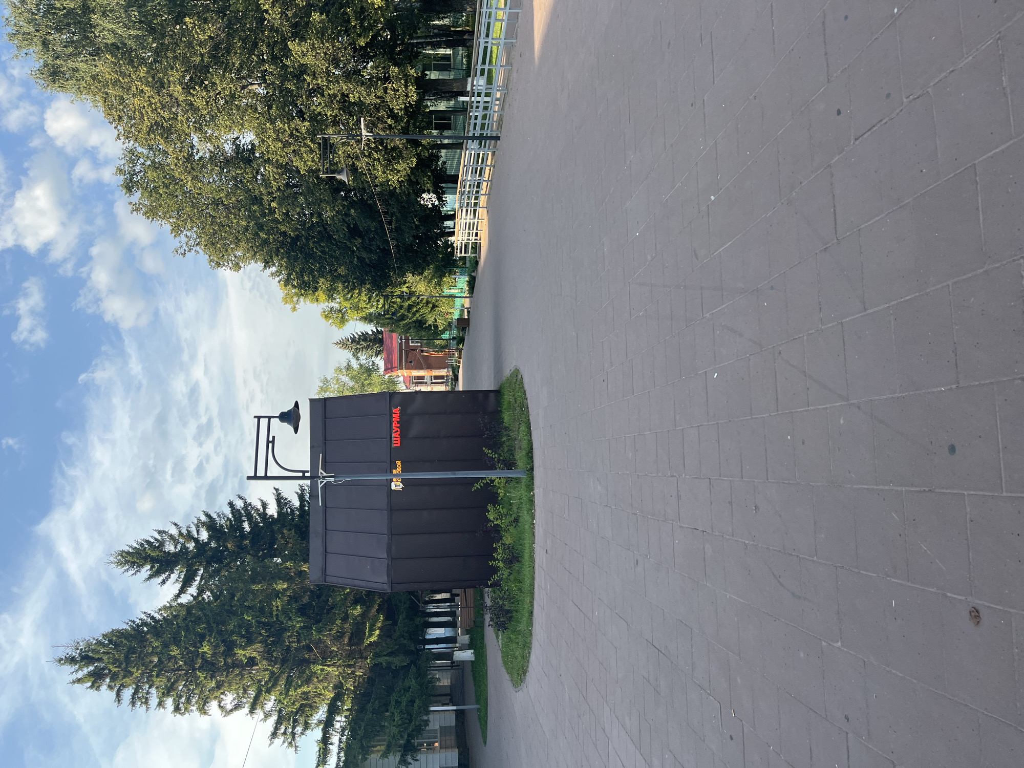 The image shows a paved area in Novye Turbasly, Russia, with a small, dark brown, cubical building in the center. The building has a sloped roof and features red neon lettering on its side that reads "ШАУРМА" (Shaurma), a type of Middle Eastern wrap. A lamppost stands in front of the building. To the left of the building are two tall evergreen trees, and beyond them, a white building with windows. To the right of the building, a white metal fence runs along the edge of a pathway, with trees visible behind it. The sky is partly cloudy with patches of blue. The ground is paved with rectangular gray tiles.