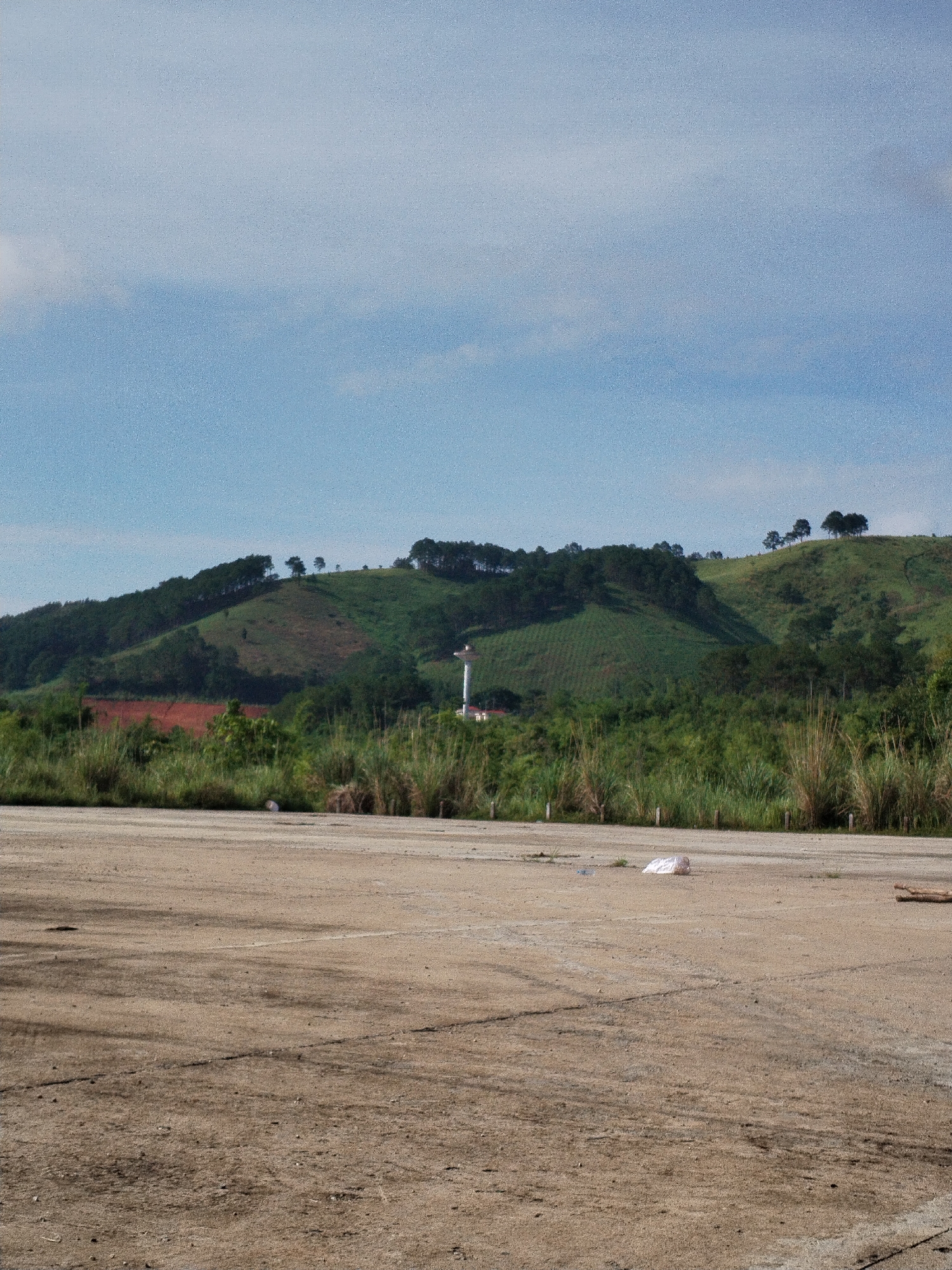 The image depicts a landscape in Bo Y, Vietnam. The foreground features a tan-colored, rough concrete surface with dirt and debris scattered across it, including a white plastic bag and small pieces of wood. In the middle ground, tall grasses and bushes separate the concrete surface from a backdrop of lush green hills. A white water tower is visible among the vegetation. The background consists of a hillside covered in trees and foliage, under a blue sky with light cloud cover. The overall scene is rural and somewhat desolate, with signs of human activity present but minimal.