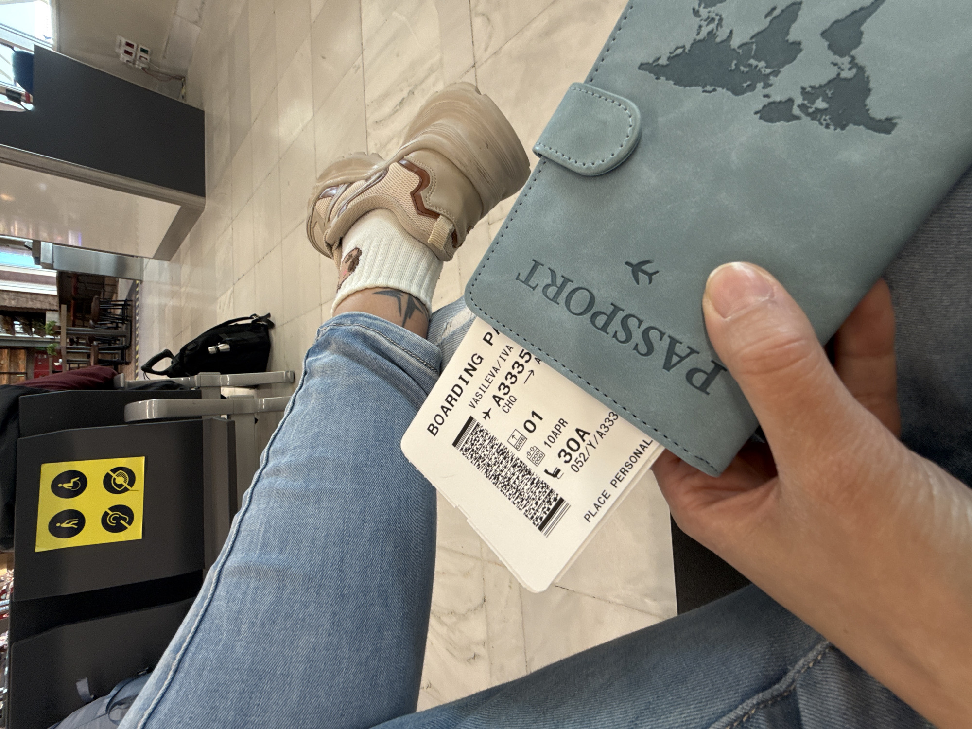 An individual is shown holding a passport and a boarding pass. The boarding pass shows "VASILEVA/IVA + A3335" and indicates "CHQ" as the airport code, seat "30A," and a date of "10APR." The person is wearing jeans and a beige sneaker with white socks. The passport cover is light blue with a world map design. The scene is set in an airport waiting area, evidenced by the presence of airport seating and luggage storage with accessibility icons on it. There is no information to determine the location in Kounoupidiana, Greece.
