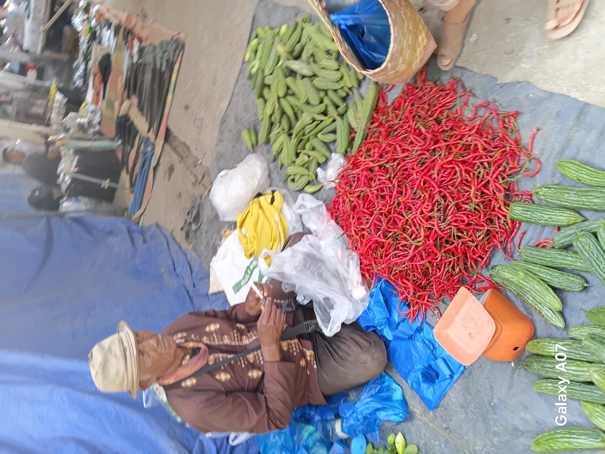 In Margo Rukun, Indonesia, an elderly Indonesian man wearing a hat and a patterned brown shirt sits on the ground beside a large pile of red chili peppers. He is holding a cigarette in his right hand and appears to be lighting it or taking a drag. In front of him, to his left, are several blue plastic bags, some containing produce. To his right, there is a small orange scale, a large pile of red chili peppers, and further to the right, a woven basket containing a blue plastic bag. Several green vegetables, possibly cucumbers or gourds, are scattered around the scene, some in front of the chili peppers and some in the background. The man is seated on a tarp, and behind him, a large blue tarp forms a backdrop. In the background, other market stalls and people are visible, suggesting a busy marketplace.