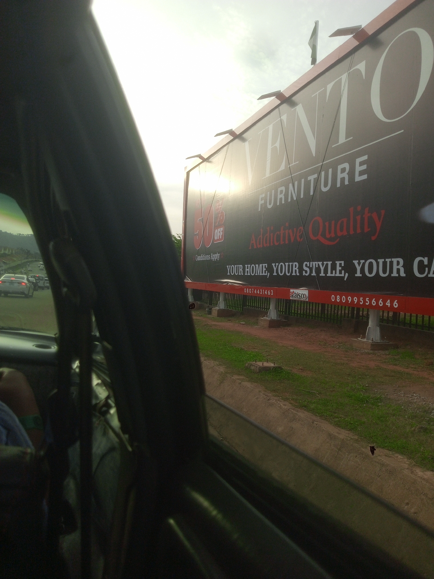 The image shows a view from inside a vehicle in Gwarinpa, Nigeria. A large billboard advertising "VENTO FURNITURE" is prominently displayed on the right side of the frame. The billboard features text such as "FURNITURE," "Addictive Quality," and "YOUR HOME, YOUR STYLE, YOUR CA." Below the main text, there are phone numbers "080744314363" and "08099556646," as well as the brand name "Kaiso." The billboard is supported by metal structures and is situated next to a grassy area and a road. In the background, there is traffic on the road, with several cars visible. The sky is overcast, with the sun partially visible. On the left side of the image, the interior of the car is partially visible, including a person's arm and shoulder wearing a blue garment with a green strap. The overall scene depicts a typical roadside view with a prominent advertisement.