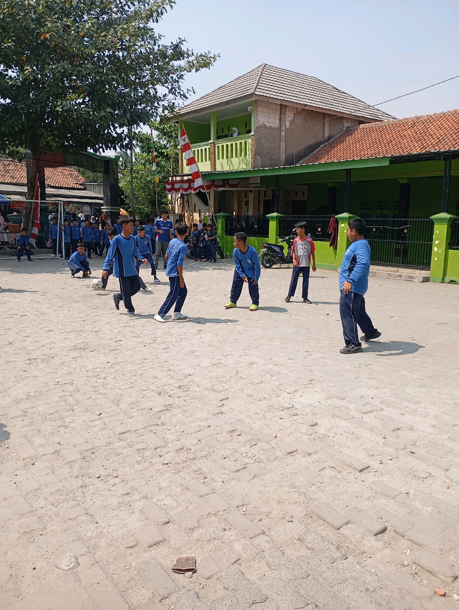 A group of young boys in blue uniforms are playing soccer on a dirt field in front of a two-story green building with a tiled roof. The building has several windows and balconies, and a red and white flag is displayed on the front. A large tree with green leaves is on the left side of the image. Several other children are watching the game from the sidelines. The scene appears to be taking place in a residential area in Babelan, Indonesia, as suggested by the style of the buildings and the presence of a motorcycle parked near the building. The ground is uneven and covered in dirt and small pebbles, with some paved sections visible. The sky is clear and blue, indicating a sunny day.