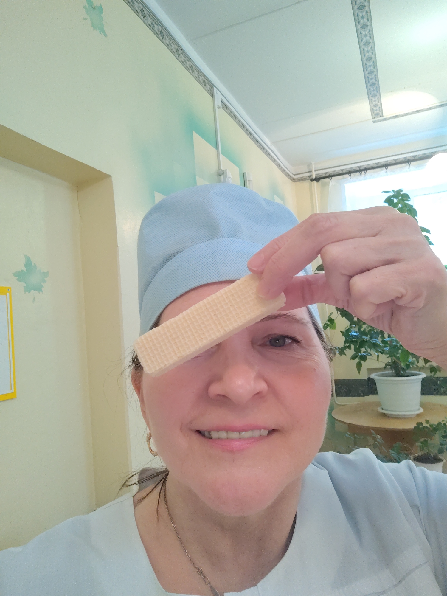 A woman, appearing to be in her 40s or 50s, is captured in a close-up selfie, smiling warmly at the camera. She is wearing a light blue scrub cap or hairnet and a light-colored, uniform-style top, accessorized with a thin chain necklace and small hoop earrings. Playfully, she holds a light-colored, rectangular wafer between her thumb and index finger, positioning it to cover her right eye.
The scene is indoors, brightly lit by natural light streaming through a window visible on the right. The room features walls painted in soft yellow and light green/blue hues, with decorative light green maple leaf shapes adorning the left wall. A small, partially visible yellow-framed sign or notice board is also present on the left. The ceiling is white and includes decorative patterned strips. In the background, a potted green plant in a white pot rests on a round wooden table near the window. The overall ambiance suggests a cheerful and lighthearted moment, possibly in an institutional or care setting in Sarapul, Russia. No specific text is legible in the image.