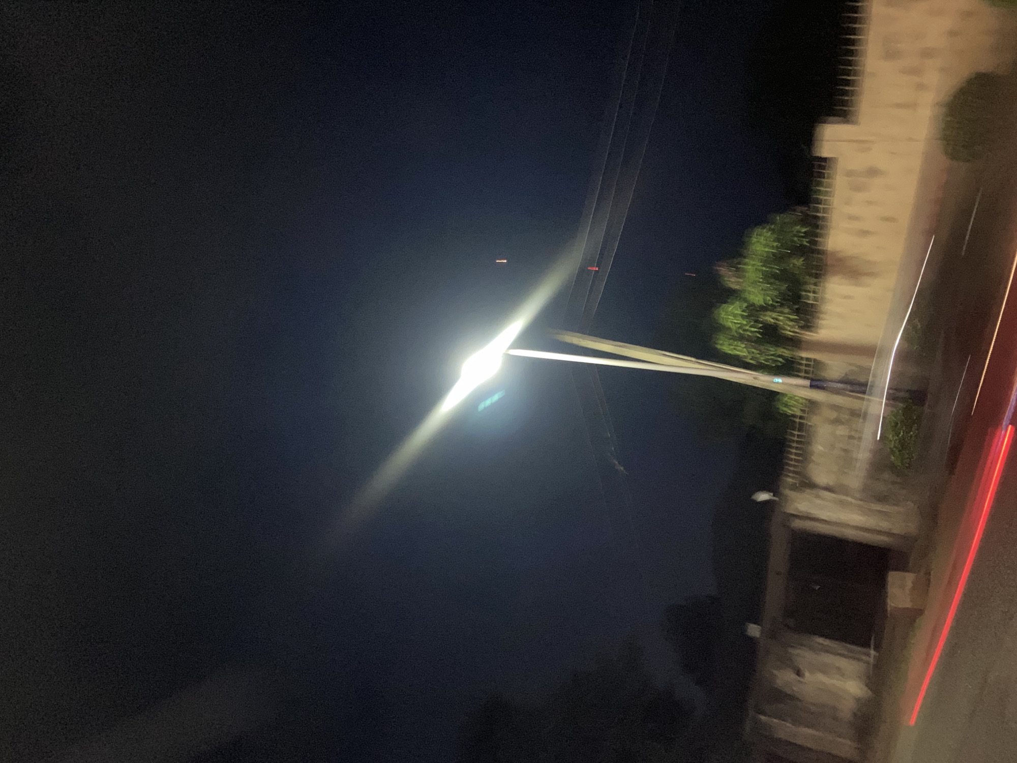 An exterior night-time shot captures a street scene in Gura Top, Nigeria. A bright street lamp illuminates a section of a road, casting a stark beam across the dark sky. Power lines are visible stretching across the frame, suspended by a pole near the lamp. The foreground shows a road with streaks of red light, suggesting moving vehicles. To the right, a low, light-colored wall borders a property, with some greenery visible behind it. A dark, recessed entrance or gate is set into a stone-like wall on the left side of the image. The overall impression is one of a quiet, possibly residential or commercial area at night.