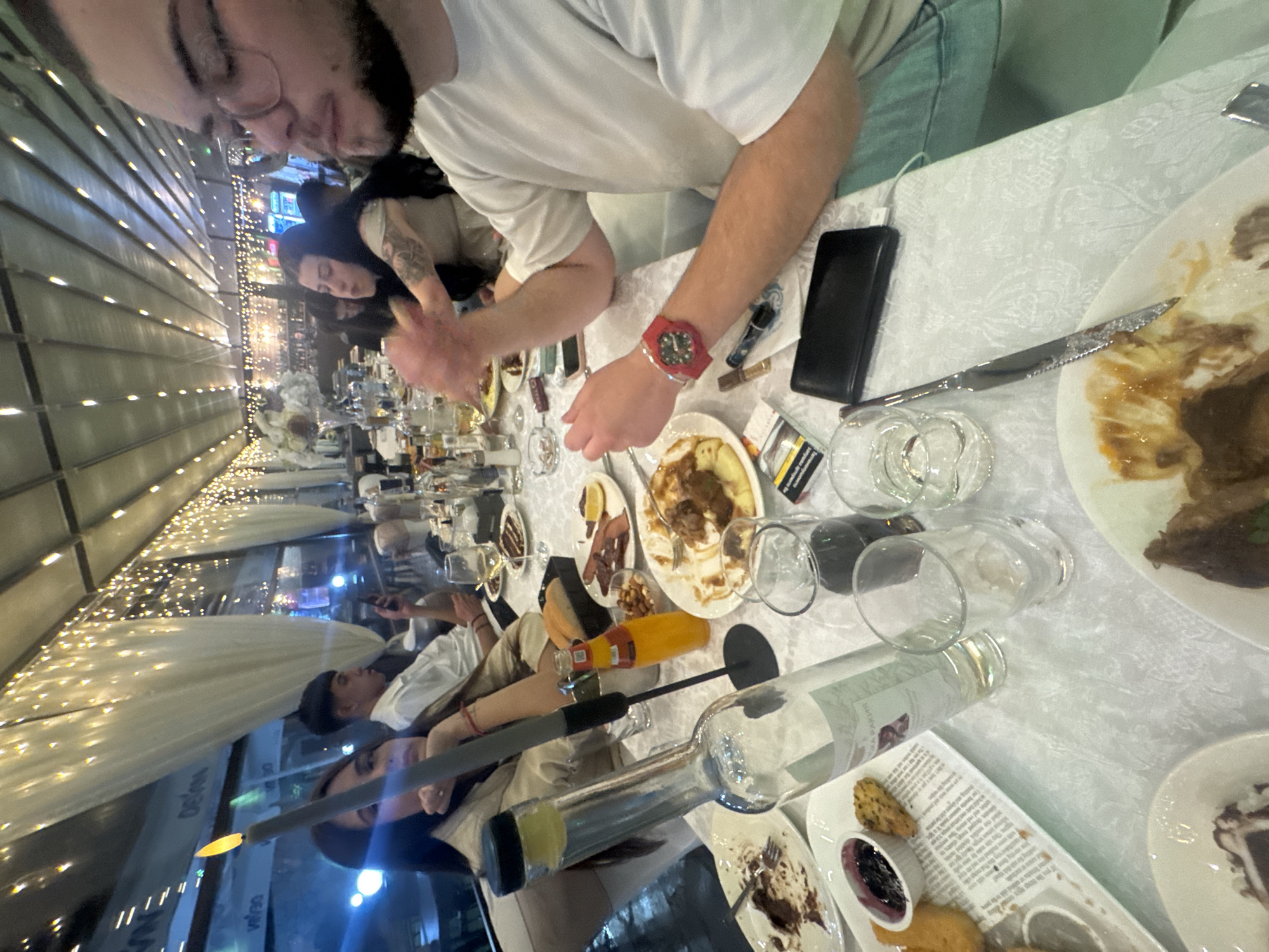 The image shows a long table at a restaurant, possibly outdoors under a retractable awning illuminated with string lights. The table is covered with a white tablecloth, and numerous people are seated along it. Food and drinks are scattered across the table, including partially eaten plates of food, empty wine bottles, glasses, and cutlery. A person wearing a white t-shirt and a red watch is prominently positioned in the foreground. Another person, wearing glasses, is positioned near the top of the image. The general setting suggests a social gathering or meal at a restaurant. I can not determine its location in Zlati Voyvoda, Bulgaria.