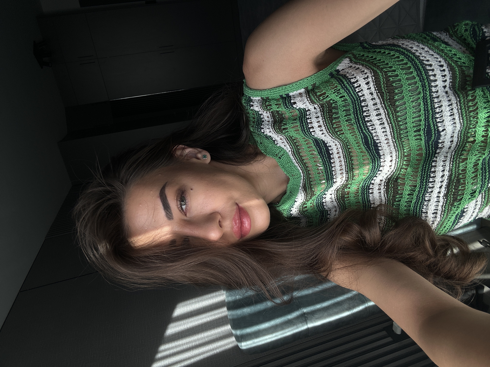 The image shows a young woman with long, brown hair, wearing a green and white striped knitted top. She is indoors, with sunlight casting shadows across her face and the left side of the image. A portion of a dark blue chair is visible on the left. The background is dark and appears to be a room with cabinetry. No specific location in Sofia, Bulgaria is identifiable from the image.