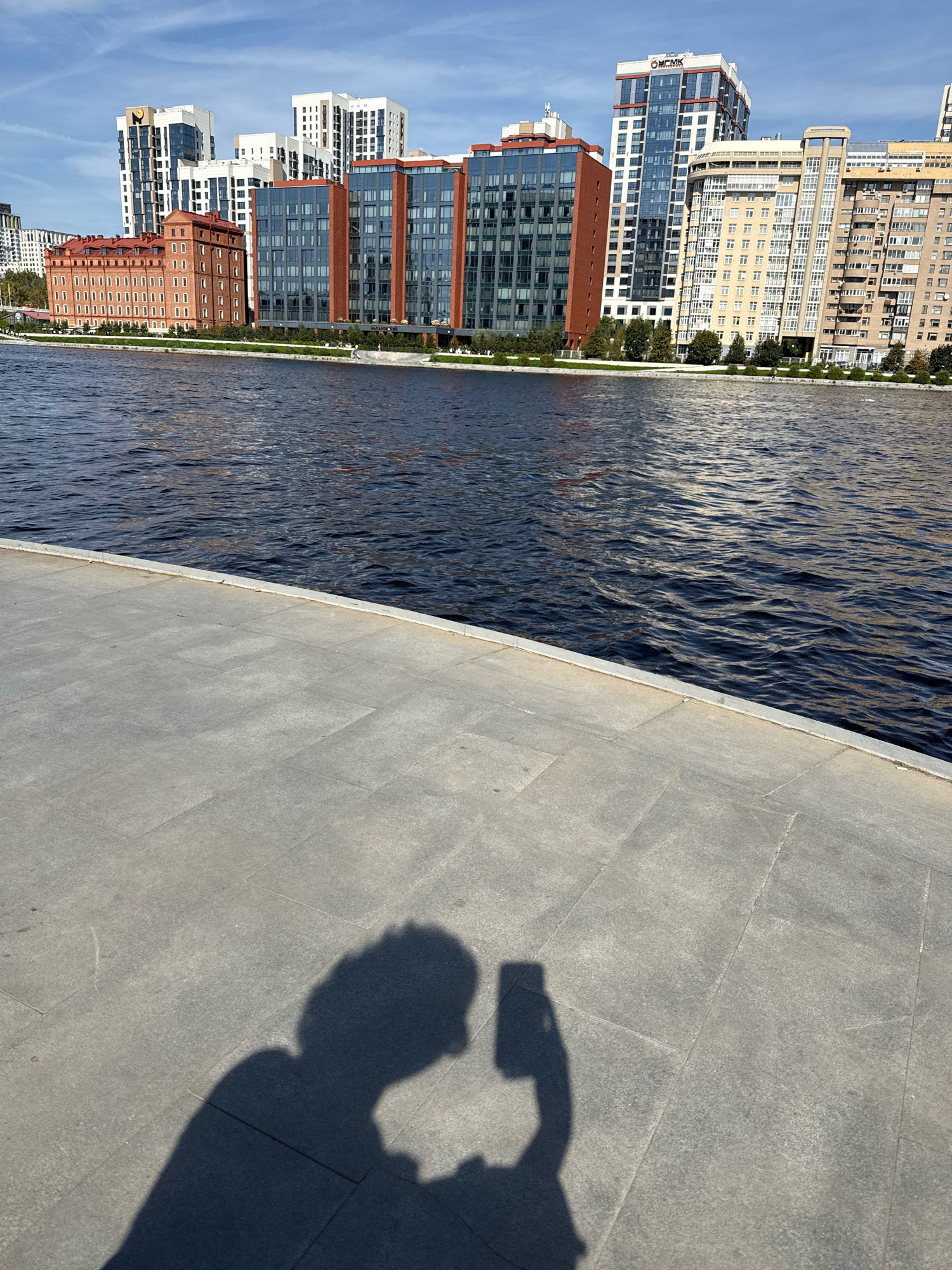 This is a full-frame, eye-level, outdoor, sunny day photograph of the Yekaterinburg City embankment near the Iset River. In the foreground, the shadow of a person holding a smartphone is cast on a grey paved surface. The person is capturing a selfie. The middle ground is dominated by the dark, rippling surface of the Iset River. In the background, a line of modern buildings stands along the embankment, including the "Onegin" residential complex, the "Summit" business center, and other residential and office buildings, showcasing a mix of architectural styles. The sky is blue with scattered white clouds.