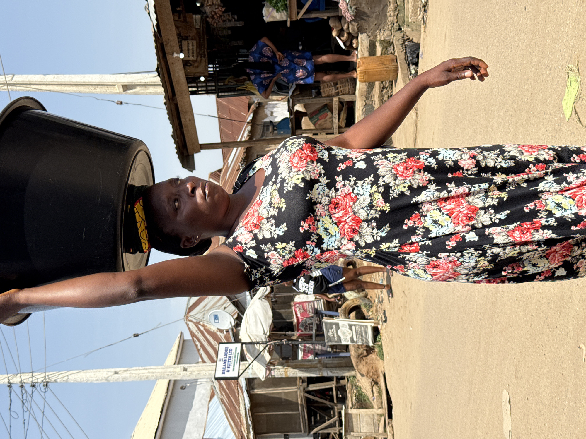 In a bustling street scene in Abuja, Nigeria, a woman carries a large black basin filled with items on her head. She is dressed in a vibrant floral print dress and walks along a dusty, unpaved road. In the background, various market stalls with corrugated iron roofs line the street, displaying goods and serving as a hub of commercial activity. Several other individuals are visible in the distance, engaged in their daily routines, contributing to the lively atmosphere of the urban environment. The day appears bright and sunny, casting clear shadows.