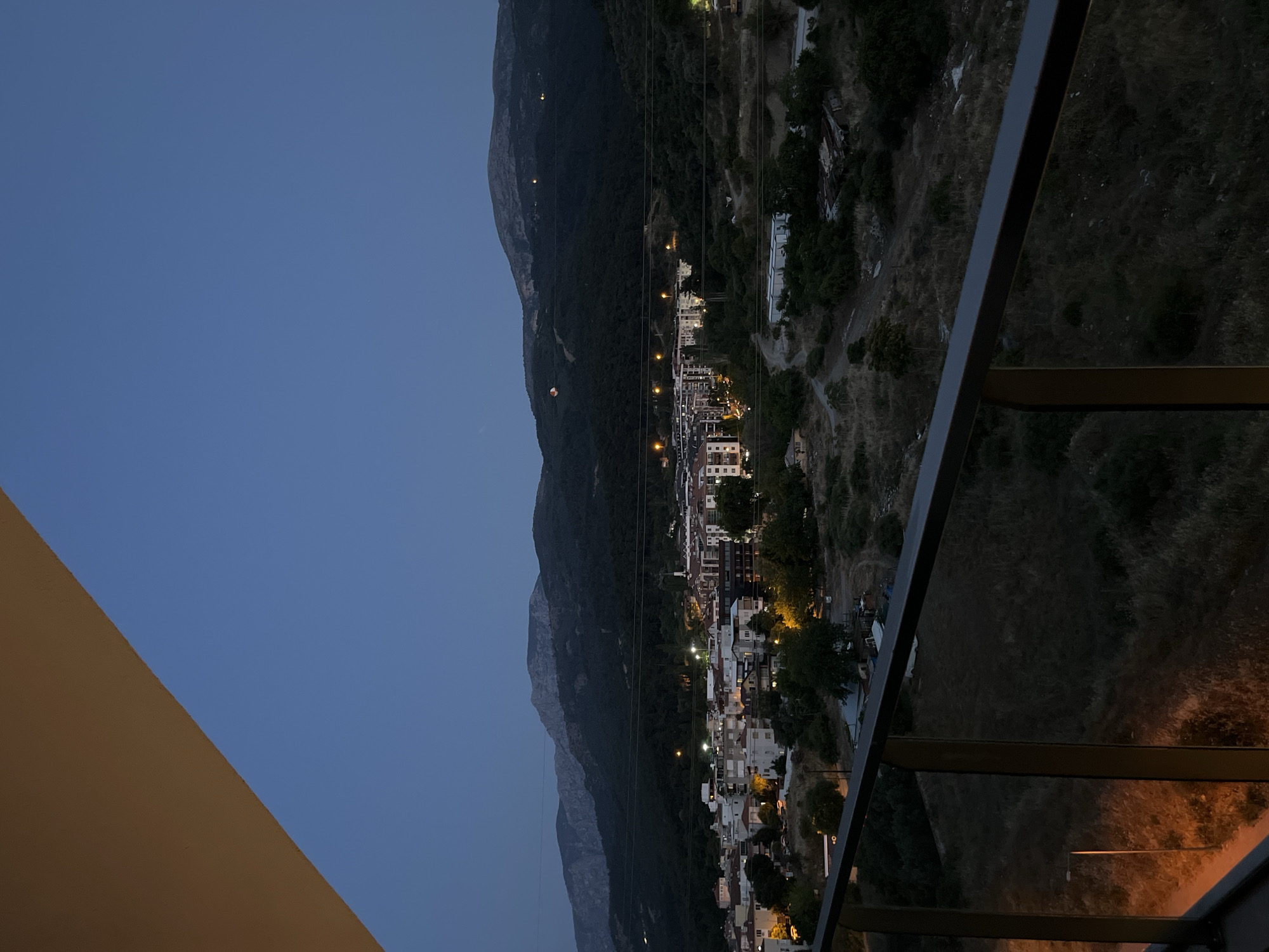 The image depicts a twilight view of a town nestled in a valley with mountains in the background. The sky is a deep blue, transitioning from lighter shades towards the horizon. In the foreground, a balcony railing with vertical supports is visible, suggesting an elevated viewpoint. Beyond the railing, the town is illuminated with streetlights and lights from buildings, showcasing a cluster of residential structures with red-tiled roofs and multi-story apartment buildings. The surrounding landscape is characterized by dry, sparse vegetation and trees. Power lines stretch across the midground, and the mountainous terrain behind the town is covered in dark, dense trees. The overall scene captures a quiet evening in a semi-urban environment, likely in a mountainous region. The specific location in Yunus Emre, Turkey, is not explicitly indicated by any visual cues within the image.