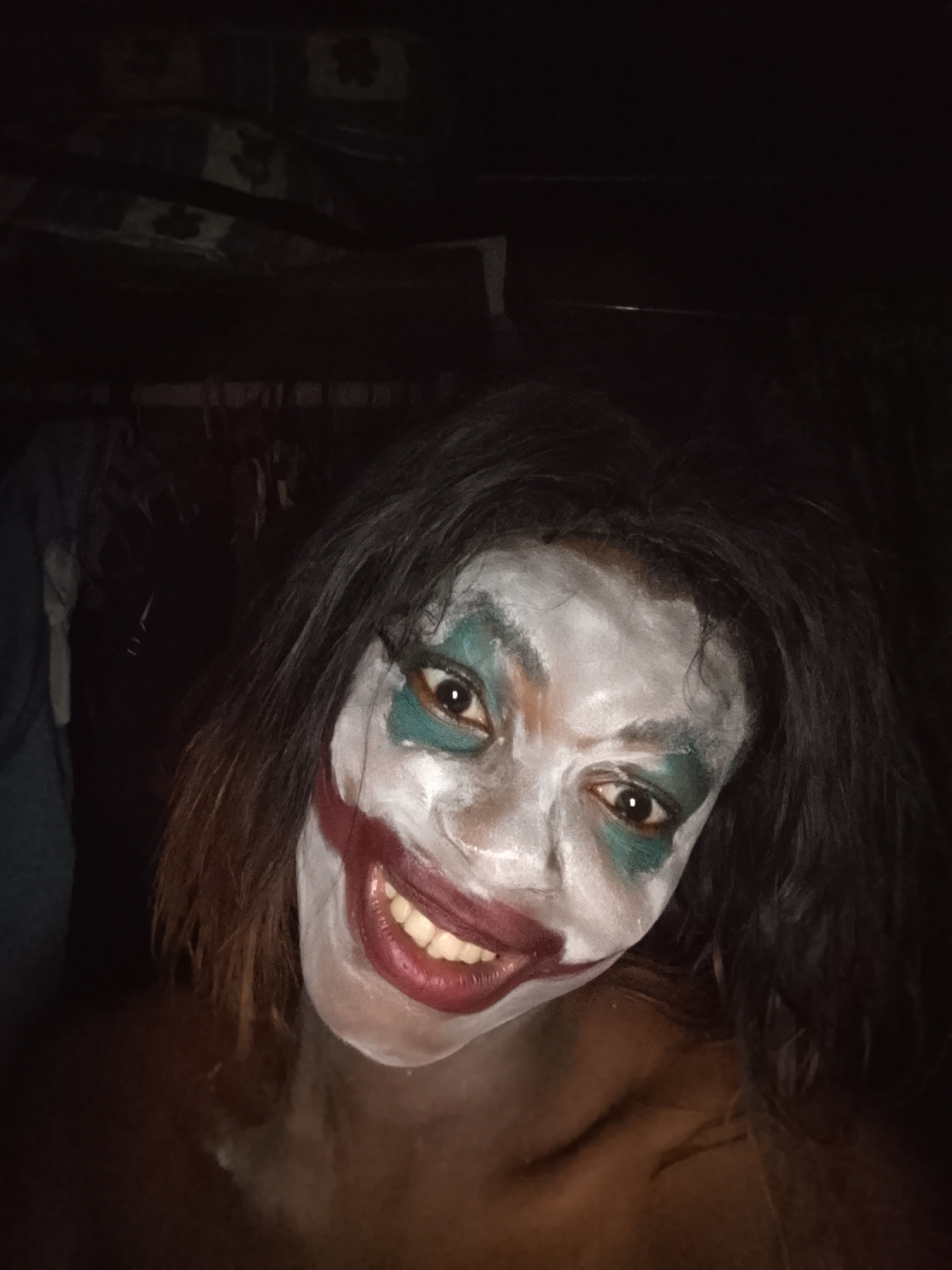A close-up, dimly lit portrait features a person wearing theatrical makeup resembling the Joker. The individual's face is painted white, with vivid green paint around their eyes and an exaggerated red smile painted across their lips, extending outwards. Their dark, messy hair frames their face, and their dark-skinned shoulder and chest are visible below the neck, suggesting bare skin or a dark top blending into the background. The person looks directly at the camera with wide, alert eyes, maintaining a prominent, unsettling grin.
The background is extremely dark and largely indistinct, with faint, shadowy patterns visible in the upper sections, possibly indicating a textured wall or draped fabric in very low light. The lighting is direct and appears to be from a single source, strongly illuminating the face and creating high contrast with the dark surroundings. The image captures a moment of posing, likely for a Halloween costume, cosplay, or performance. There is no visible text in the image.
