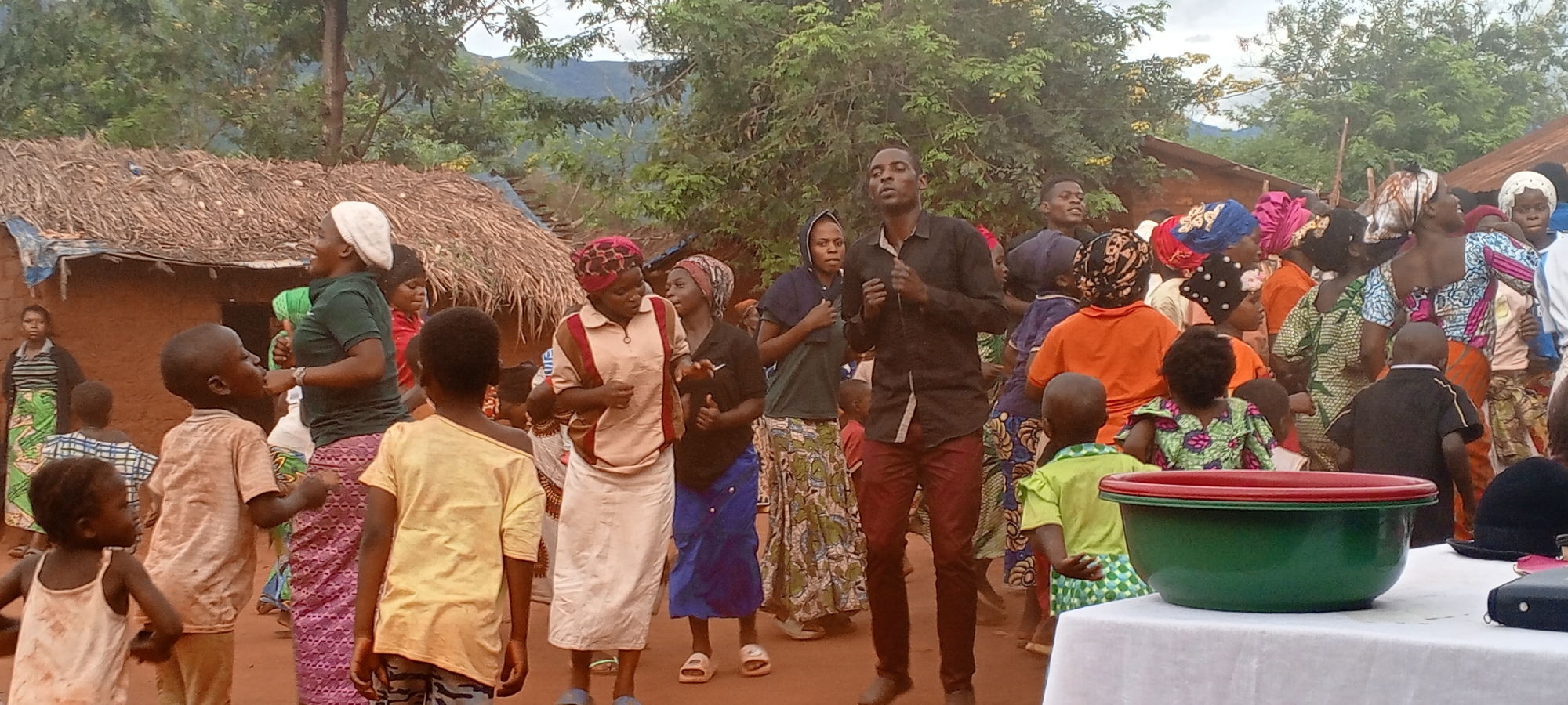 The image shows a gathering of people in what appears to be a rural or village setting in Ngalula, Democratic Republic of the Congo. A group of adults and children are positioned outside of simple dwellings constructed with mud walls and thatched roofs. Many individuals are dressed in colorful, patterned fabrics and head coverings, typical of local dress. A man in a dark shirt and reddish trousers is prominently placed in the center of the gathering. Some people appear to be dancing or moving rhythmically. A table with a large green and red bowl is visible in the foreground. The background includes trees and what seem to be distant hills.