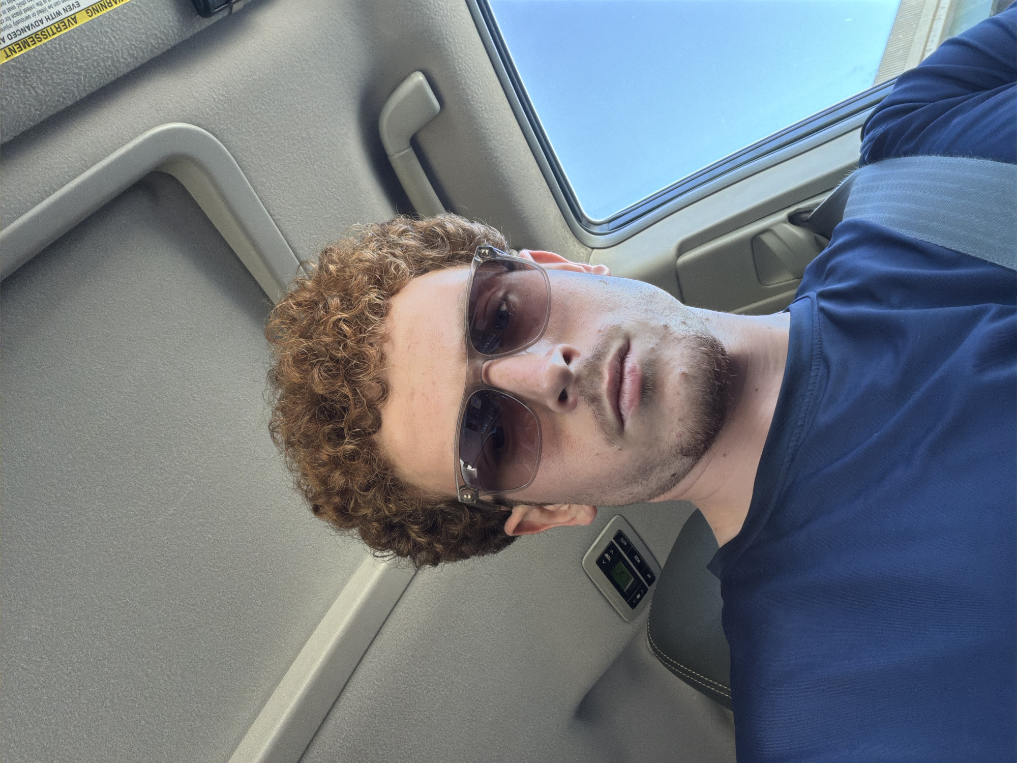 A male with curly auburn hair and a goatee is looking at the camera while sitting in a car. He is wearing sunglasses and a navy blue shirt. The car has a sunroof open to a clear blue sky. There is a yellow warning label on the sun visor. The image was taken in Pepper Pike, United States of America.