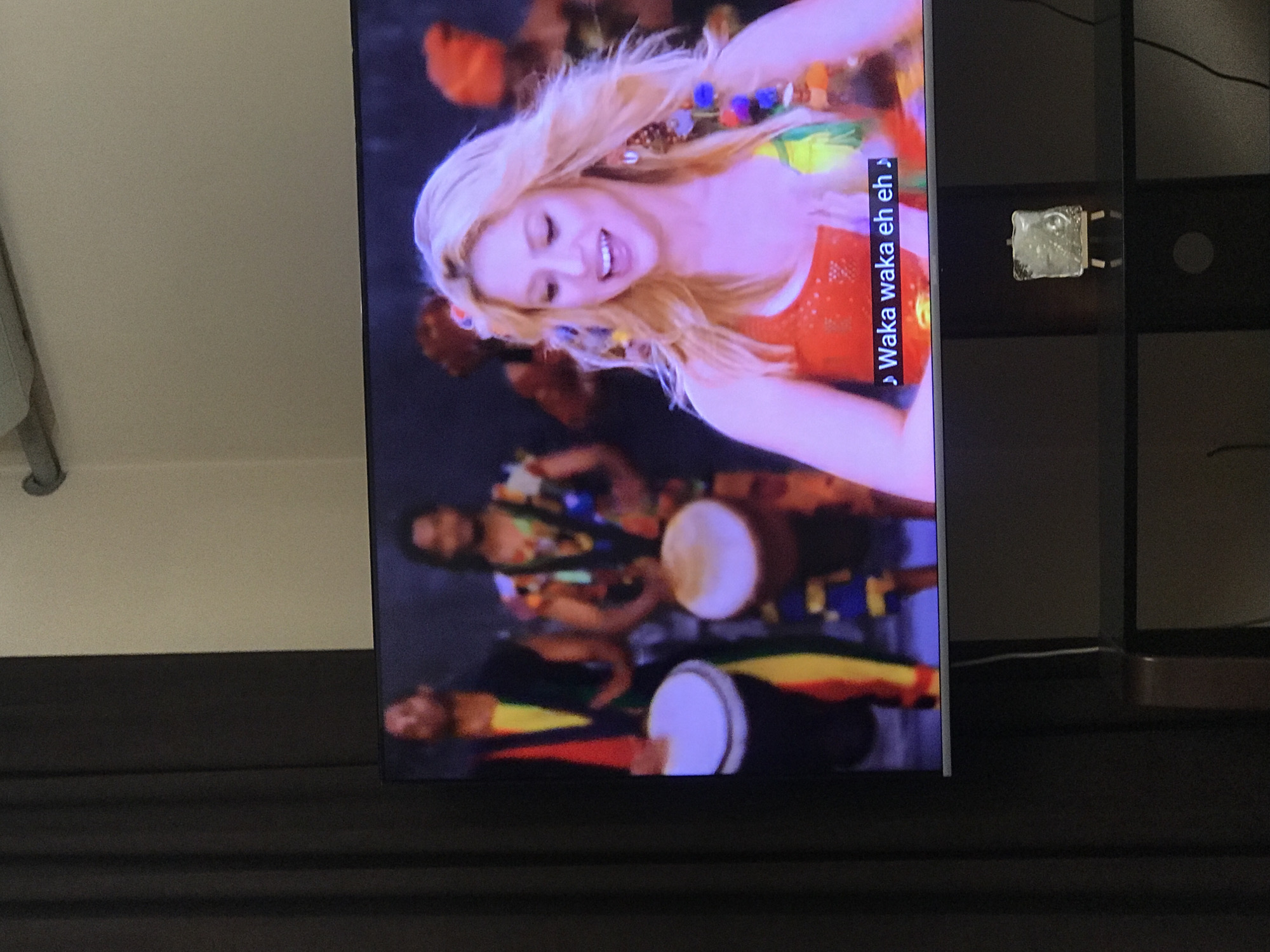 The image shows a television screen displaying a scene with Shakira singing her song "Waka Waka". There are other people in the background playing drums. The text on the screen says "Waka waka eh eh". The TV is on a dark-colored stand with a small decorative object on a shelf above it. The background shows a portion of a white wall. There is no information in the image to determine location in Yerevan, Armenia, or if it is even located in that city.