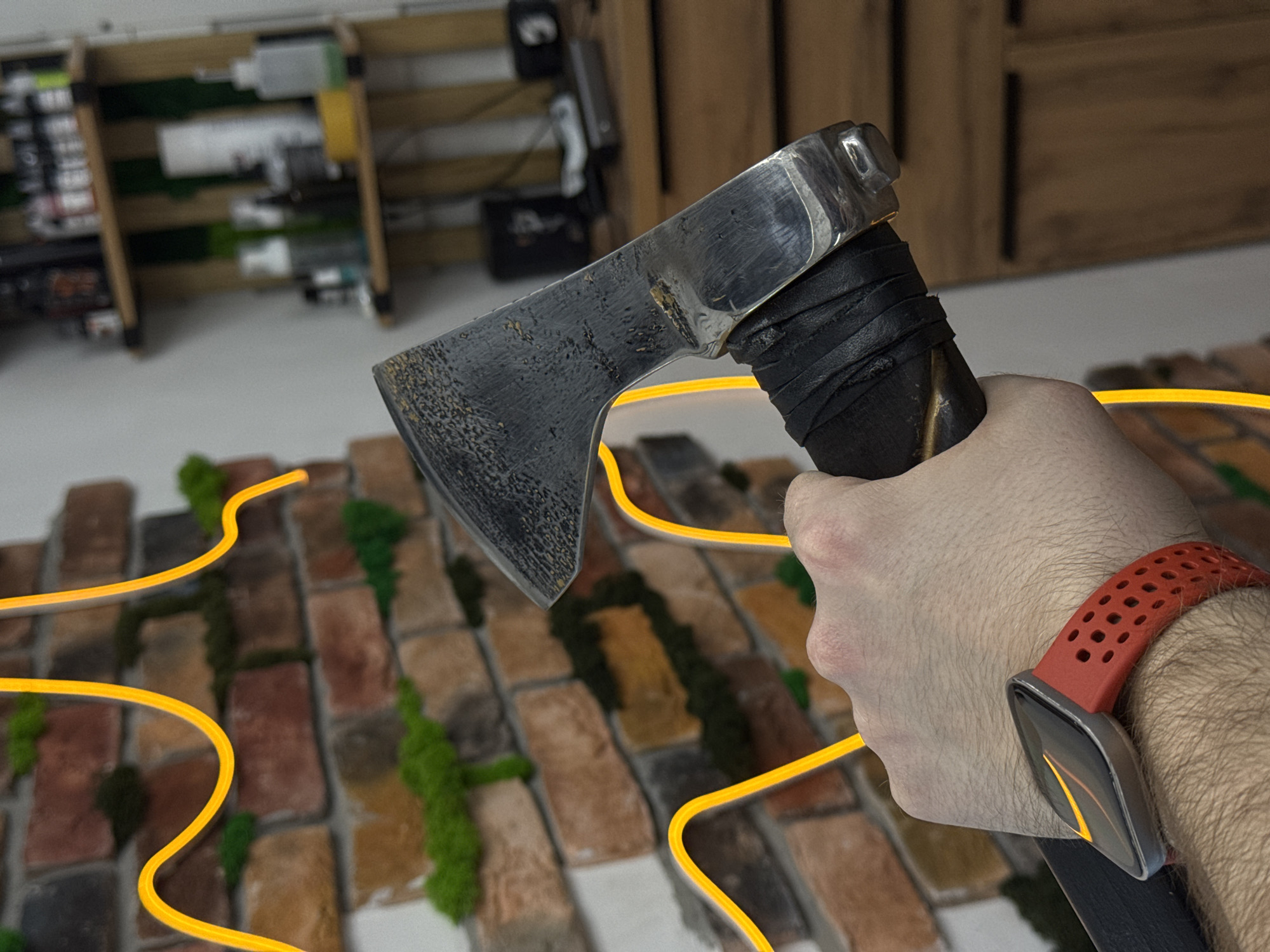 A close-up shot depicts a hand holding a small axe. The axe has a dark, weathered blade and a leather-wrapped handle. The hand is fair-skinned and wears a red and black smartwatch on the wrist. The background consists of a textured wall made of bricks and patches of green moss, with a faint yellow light trail meandering across the wall. In the blurred background, a wooden shelf unit containing various items is partially visible. The location or city of Chaikovsky, Russia, cannot be determined from the visual information provided.