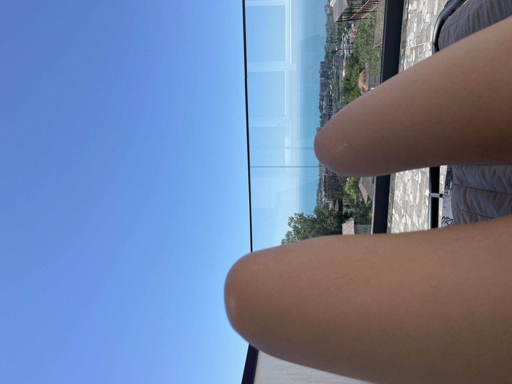 The image captures a first-person perspective of a person lounging outdoors on a clear, sunny day in Baranovka, Russia. In the immediate foreground, the upper thighs and knees of two tanned human legs are visible, resting on a grey cushioned lounger. A modern glass balcony railing with a dark top frame extends horizontally across the midground, reflecting portions of the sky. Beyond the railing, a sprawling coastal city with diverse architecture and abundant greenery descends towards a large body of calm blue water, presumably the Black Sea, which meets the horizon. The upper half of the image is dominated by an uninterrupted, vivid blue sky.