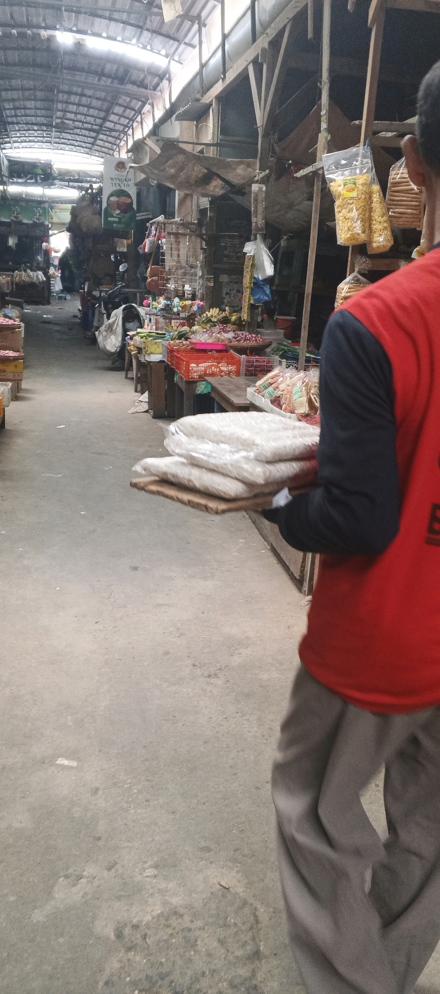 The image shows a market scene in Sekaran, Indonesia. A person wearing a red shirt and gray pants is walking away from the camera, carrying a flat tray stacked with several white, rectangular packages. The market is covered by a corrugated metal roof supported by wooden beams. Stalls line a concrete walkway, displaying various goods including produce like bananas and red onions, packaged snacks, and other items. The overall atmosphere suggests a bustling marketplace.
