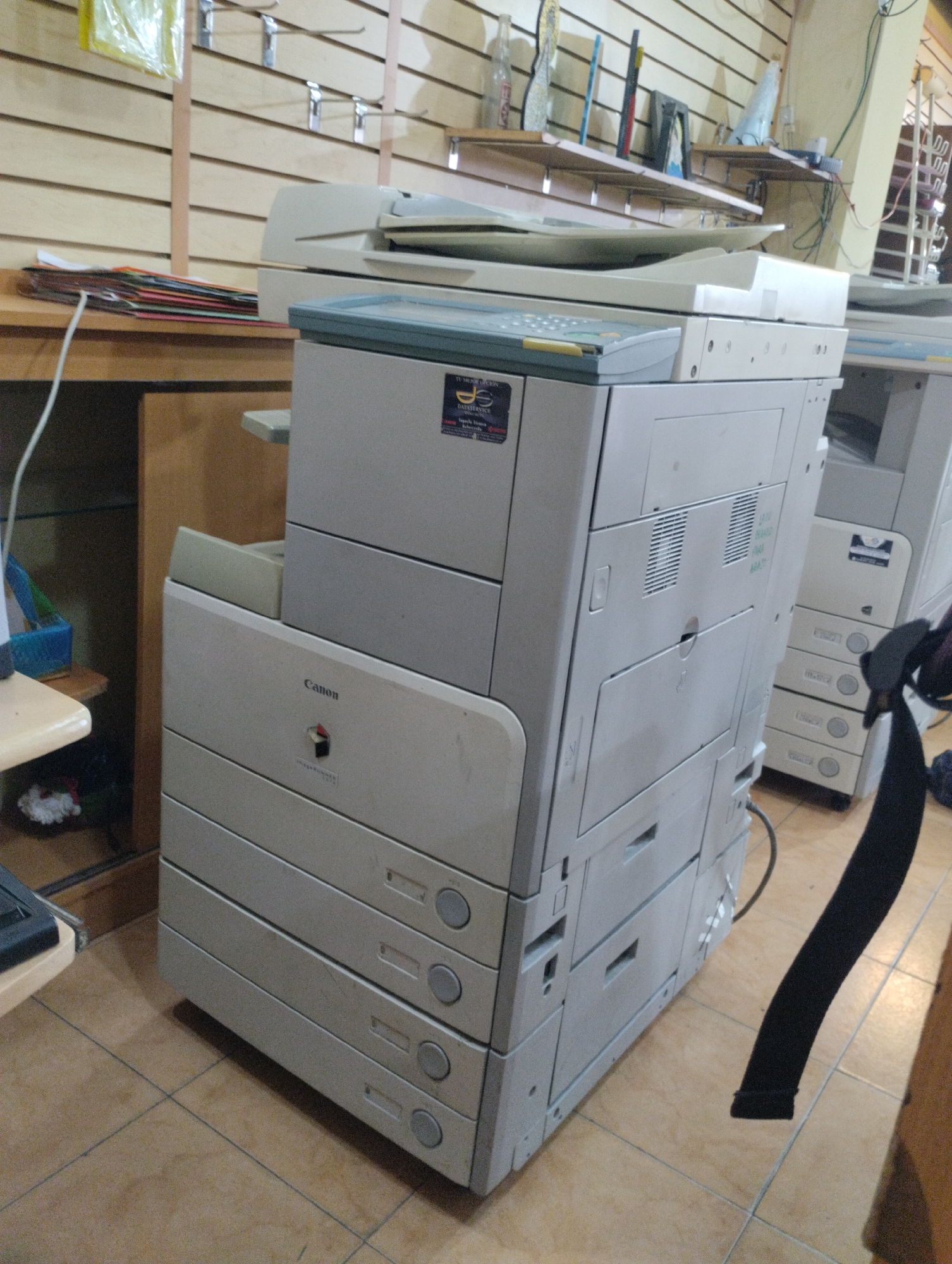 The image presents an indoor commercial or office environment located in Juan Griego, Venezuela. The central focus is a large, light grey Canon imageRUNNER multi-function printer (MFP), positioned in the mid-ground. This unit comprises a flatbed scanner/document feeder assembly on top, featuring a control panel with yellow and grey push-buttons. Below this, the unit is stacked with at least four distinct paper input trays. A black sticker with "DATASERVICE" text is visible on the upper left side of the MFP's main body.
To the right and slightly behind the primary MFP, a second, similar light grey MFP with multiple paper trays is partially visible. A grey electrical cable extends from the rear of the main MFP, running along the tiled floor.
The background wall is covered with light-toned wooden slatwall paneling, equipped with several chrome-finished display hooks. A light brown wooden desk or counter is situated to the left of the main MFP, supporting a stack of colorful folders. Further left and lower, another small wooden shelf unit holds a blue plastic basket. Above the secondary MFP, wooden shelves contain various indistinct objects, including what appears to be bottles and other supplies. A white cable descends from the left desk area towards the floor.
The floor is covered in light brown square ceramic tiles with clearly visible grout lines. A black strap-like object hangs vertically near the right side of the main MFP, extending down to the floor. No individuals are present within the frame, and no actions or interactions are depicted. The scene suggests a functional area, possibly a copy center, office supply store, or an administrative workspace.