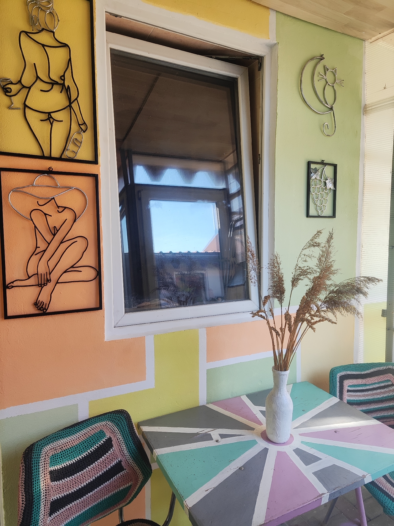 The image depicts a colorful outdoor seating area on a porch or patio, likely in Gvardeysk, Russia, given the prompt's context. In the foreground, a square table with a geometric pattern of light blue, pink, grey, and white lines is visible. On the table, a white vase holds a bouquet of dried grass or reeds. To the left of the table, a chair is covered with a crocheted seat cover featuring stripes of black, green, pink, and white. Another similar chair is partially visible to the right of the table.
The wall behind the table is painted in a patchwork of pastel colors: peach, yellow, light green, and white. Two pieces of metal wall art are mounted on the peach-colored section of the wall. The upper artwork is a black wire silhouette of a nude female figure holding a wine glass and a bottle. Below it is another black wire silhouette of a nude female figure wearing a wide-brimmed hat, seated with her legs crossed.
A white-framed window is positioned centrally in the composition, above the table and artwork. The window is open, revealing the sky and some rooftops outside. To the right of the window, on the light green wall, is another piece of metal art depicting a stylized cat sitting on a crescent moon. Below the cat artwork is a small, dark frame containing a depiction of grapes. The ceiling of the porch appears to be made of light-colored wood planks. The overall impression is of a decorated, possibly bohemian or eclectic, outdoor relaxation space.