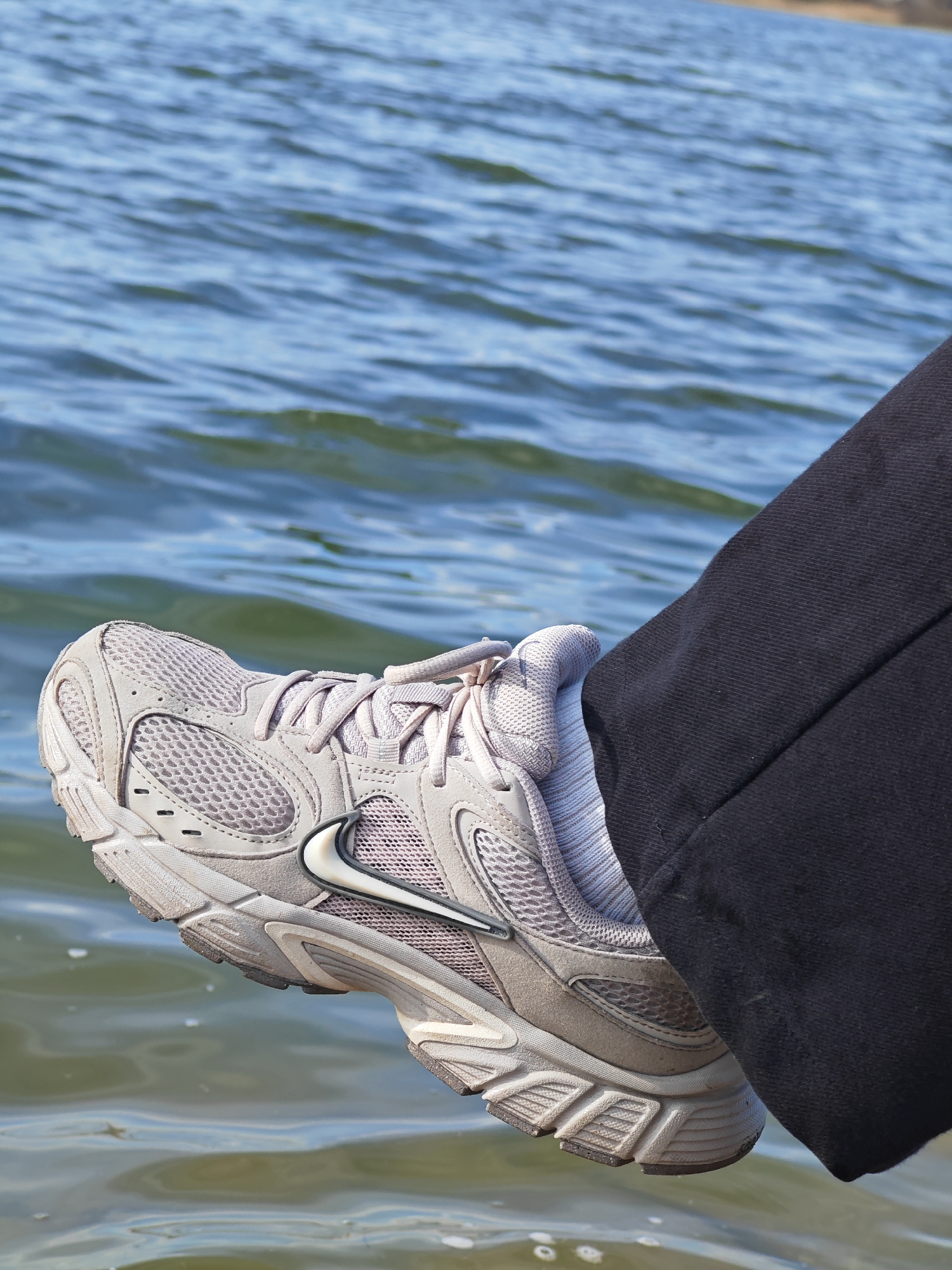 A close-up shot shows a leg with a gray Nike sneaker extending over a body of water. The leg is clad in dark pants. The background is dominated by rippling blue water. There is no indication that the scene is located in Shumen, Bulgaria. There are no people visible other than the leg in the shot.