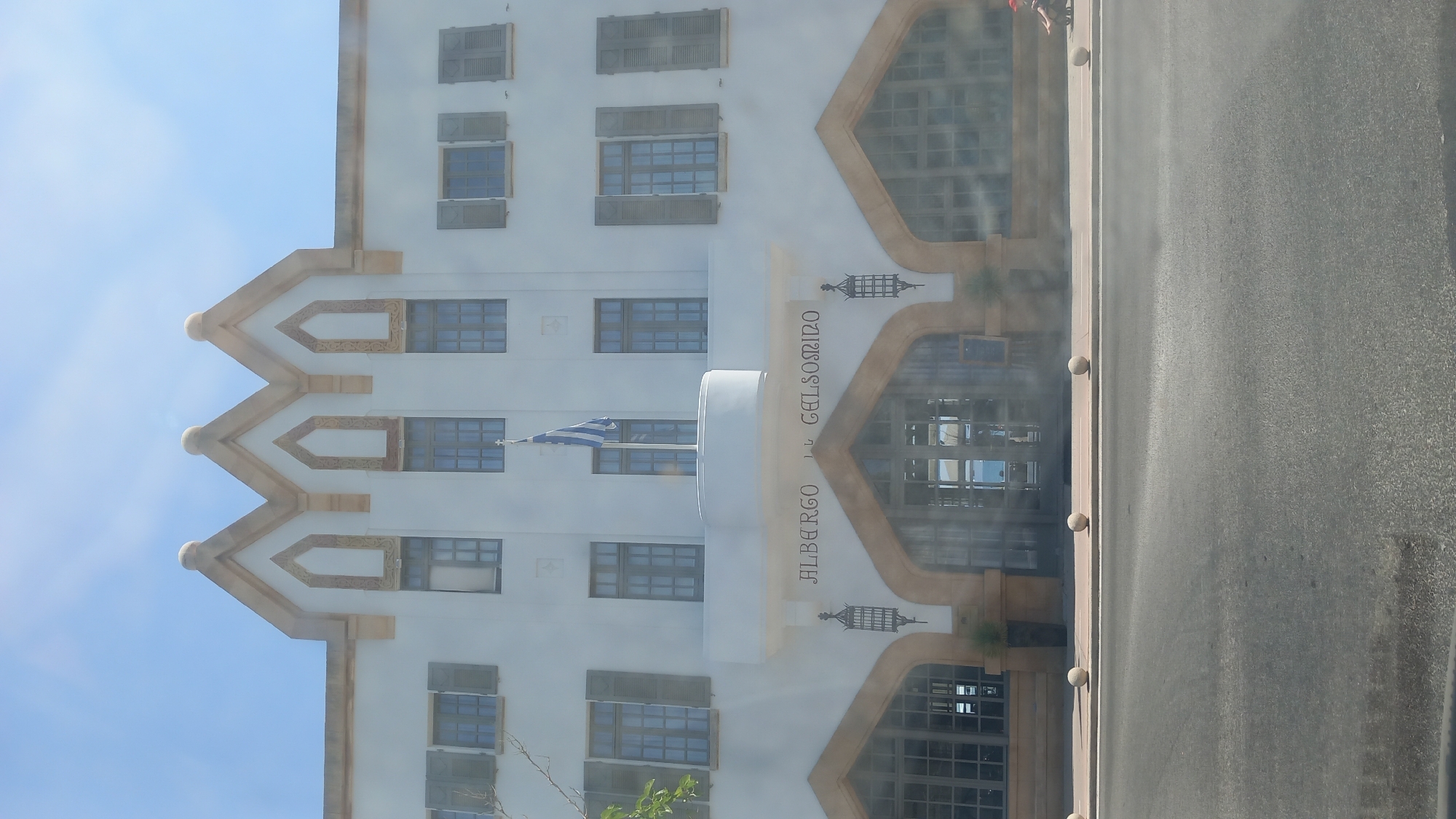 The image shows the facade of a white building with arched entrances and windows, decorated with a light brown trim. The building has a distinctly architectural style, with a triple-peaked roofline adorned with decorative elements. On the central upper section of the building, the words "ALBERGO DEL GELSOMINO" are visible. A blue and white flag, likely the Greek flag, is flying from a pole on a balcony. The building is situated on a street, with a road in the foreground and several spherical bollards lining the sidewalk. The sky is clear and blue, suggesting a sunny day. A person is visible on the right side of the image, seated near the building. Based on the sign, the location is Albergo del Gelsomino in the city of Kos, Greece.