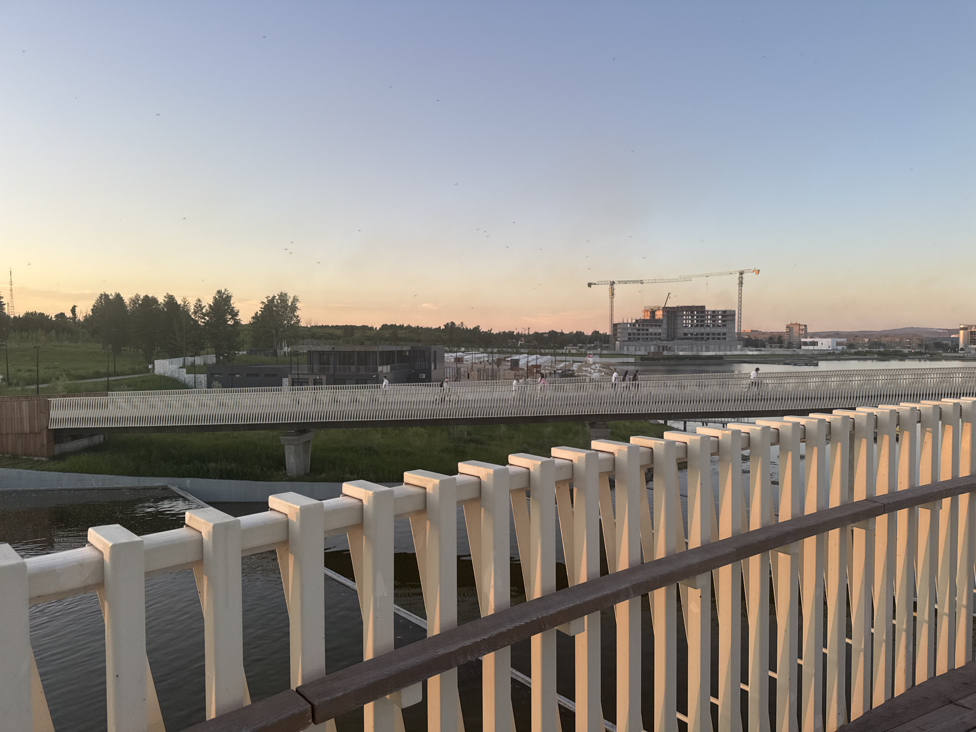 The image depicts a pedestrian bridge crossing a body of water, likely a river or canal, in Magnitogorsk, Russia. The bridge features a white, slatted railing in the foreground, with wooden and white elements forming its structure. In the background, another pedestrian bridge stretches across the scene, with several people visible walking on it. Beyond the bridges, a developing cityscape is apparent, with several multi-story buildings under construction, including a prominent structure with cranes. The sky displays a gradient of colors, transitioning from a pale orange and yellow near the horizon to a light blue overhead, suggesting either sunrise or sunset. Trees and green spaces are visible along the riverbanks, creating a contrast between nature and urban development.