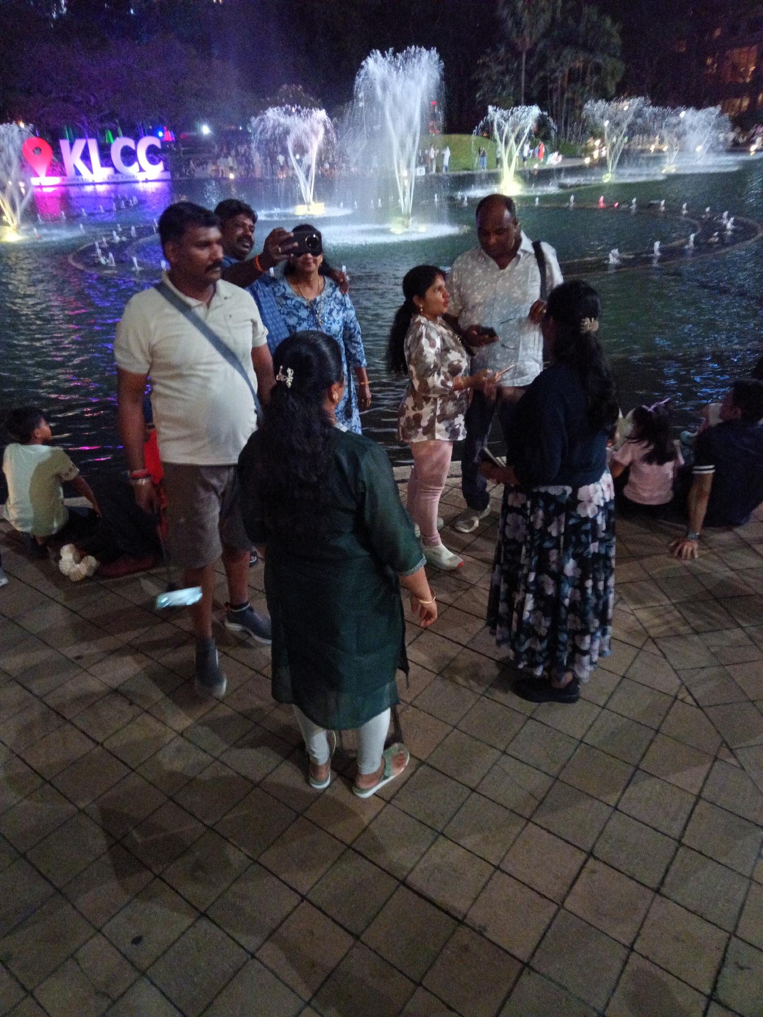 The image depicts an outdoor scene at night, likely at the KLCC Park in Kuala Lumpur, Malaysia, given the prominent "KLCC" signage illuminated in pink and blue. Several individuals are present, most of whom appear to be tourists enjoying the fountain display.
In the foreground, a man wearing a light-colored polo shirt and grey shorts stands with a blue strap across his chest. To his right, a woman in a blue patterned top and a dark skirt is holding a mobile phone, possibly taking a picture or recording a video. Behind them, another man with a darker complexion and a patterned shirt is looking towards the camera and also holding a phone. Further to the right, a woman in a dark top and a floral skirt is also present.
In the mid-ground, a child in a light-colored t-shirt is seated on the ground near the water. Behind the main group, a fountain sprays water upwards, illuminated by lights. Additional fountains and water features are visible in the background, as well as palm trees and other greenery, suggesting a park setting. The ground is paved with square tiles. The overall atmosphere appears to be relaxed and festive, with people enjoying the evening ambiance.