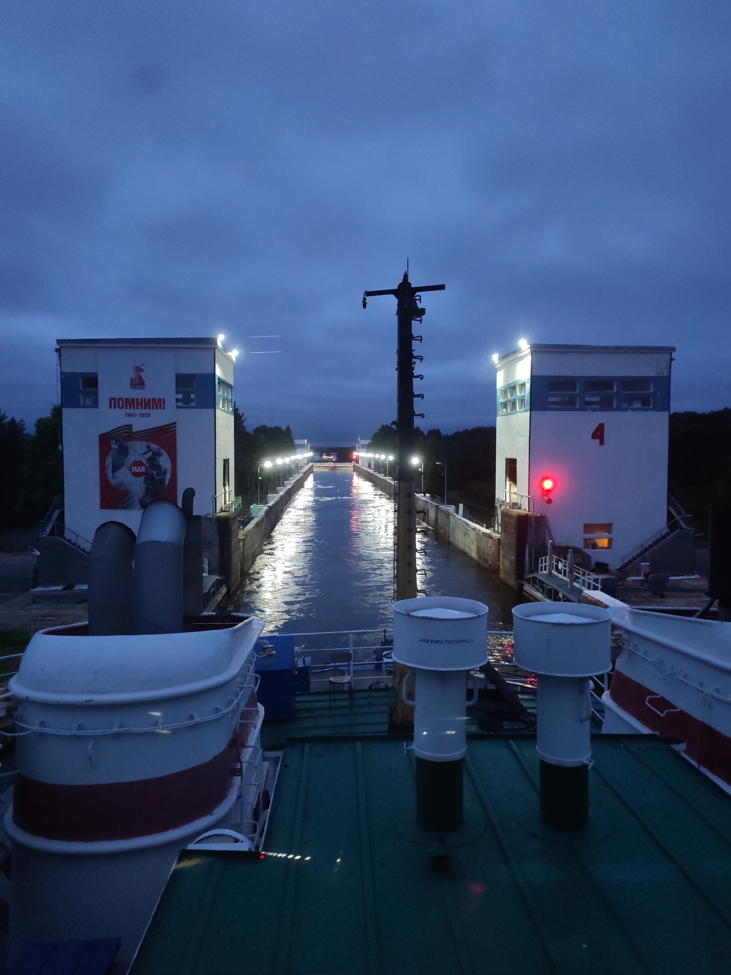 The image shows a view from a ship's deck at dusk, looking down a canal towards a lock system in Markovo, Russia. Two identical, white, multi-story buildings flank the canal, illuminated by overhead lights. The building on the left has a large red and white mural featuring a circular image with figures and the word "МАЯ" (May) in Cyrillic script, along with the text "ПОМНИМ!" (We Remember!) and dates. The building on the right is marked with the number "4" in red and a red signal light. The canal reflects the lights from the buildings and the sky, creating a shimmering effect. In the foreground, parts of the ship are visible, including white structures with red banding and two cylindrical white vents. The sky is a deep blue, indicating twilight or a cloudy day.