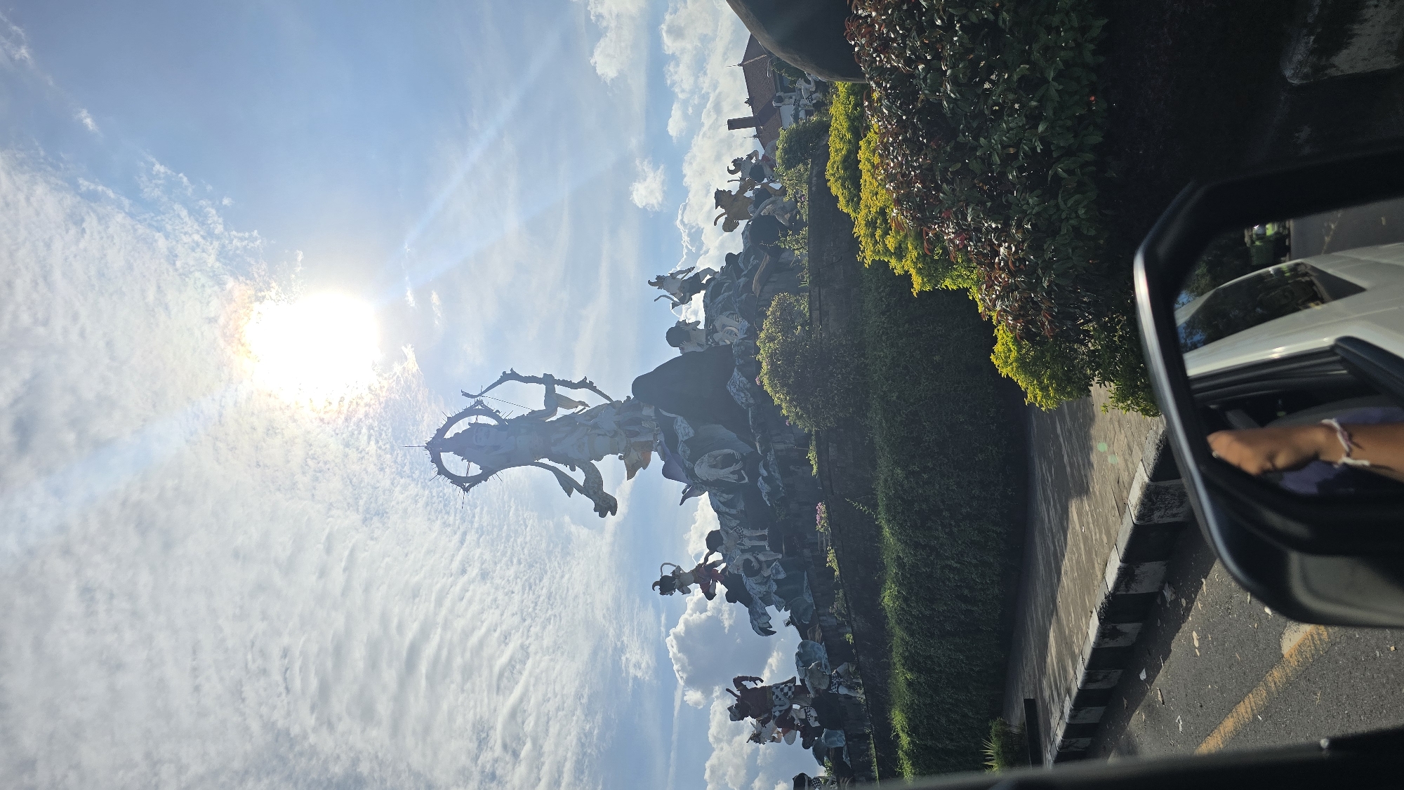 The image captures the Arjuna Wijaya statue in Denpasar, Indonesia, under a bright, slightly obscured sun against a partly cloudy sky. The statue features figures in a dynamic scene, with a central archer figure. Lush greenery is visible to the right of the statue. A car window is in the foreground, with a partial view of a person's hand and arm, indicating the photo was taken from inside a vehicle. A road is visible beneath the greenery.