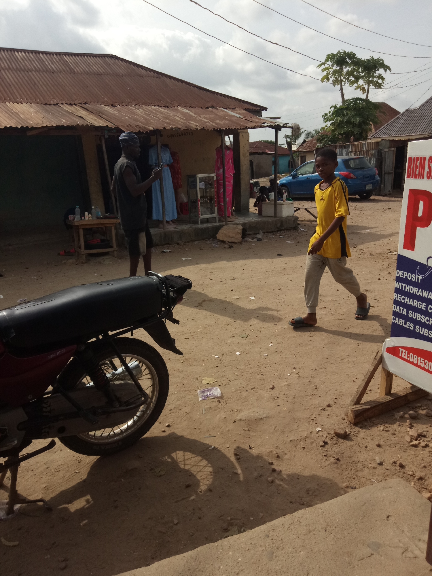 The image depicts an outdoor scene in what appears to be a village or a less developed urban area. In the foreground, a red and black motorcycle is parked, casting a shadow on the dusty ground. A young boy wearing a yellow t-shirt with black stripes and grey pants is walking from right to left, his gaze directed towards the viewer. Behind him, a blue sedan is parked on the dirt road.
In the middle ground, a man wearing a dark sleeveless shirt and a blue head covering is standing and looking at his phone. He is next to a stall with a rusty corrugated metal roof, displaying several items of clothing, including blue and red dresses. A wooden table with bottles and other small items is also present.
Further in the background, more buildings with corrugated metal roofs and various structures are visible, suggesting a cluster of small businesses or homes. A large white sign with red lettering, partially obscured, stands on the right side of the image, with visible text including "BIEM S" and "TEL:081530". The ground is predominantly dirt and sand, with scattered debris. The sky is cloudy.