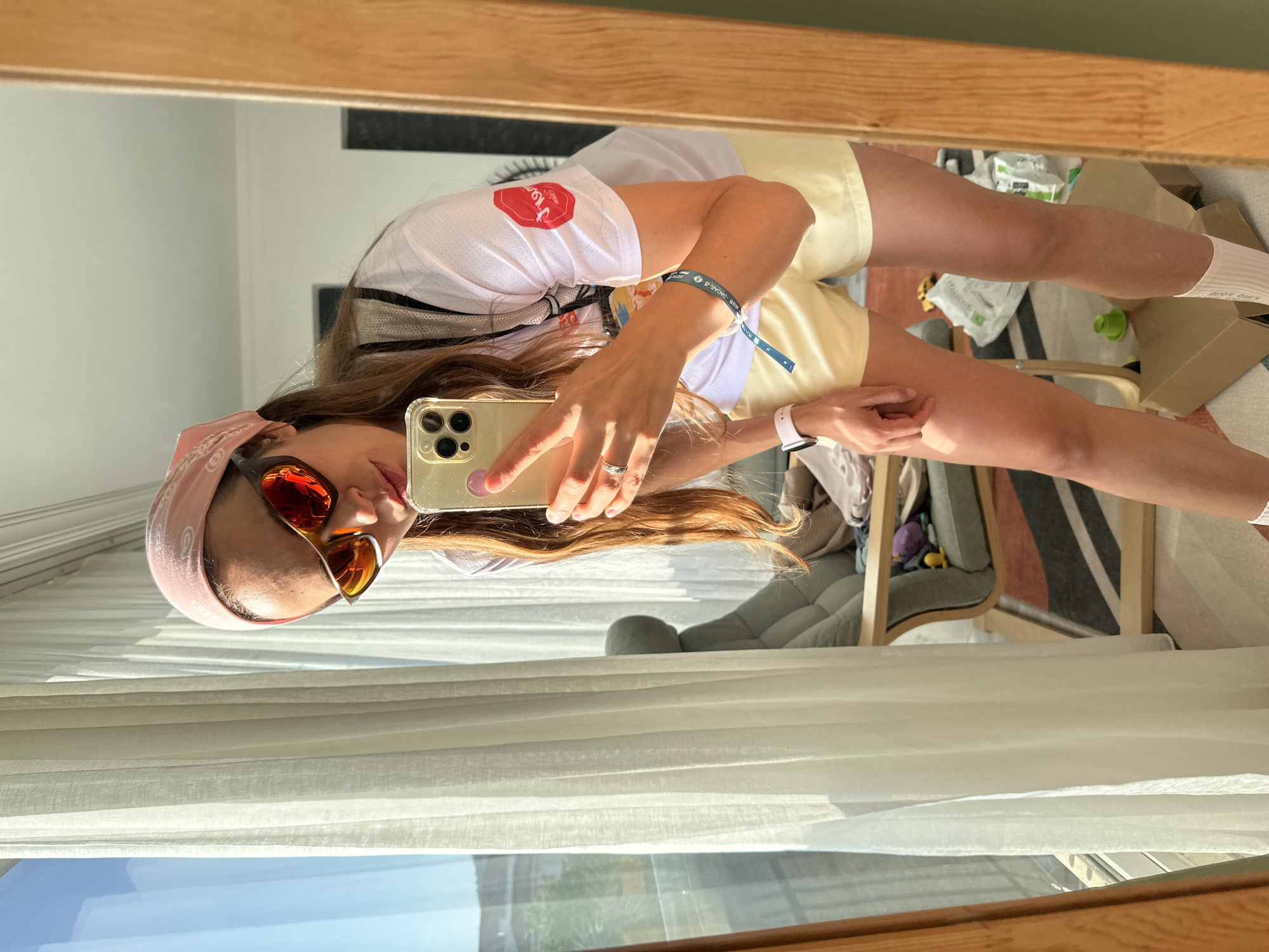 A woman is taking a selfie in a mirror. She is wearing a pink bandana, orange sunglasses, a white shirt with a red logo on the sleeve, yellow shorts, a blue bracelet on her left wrist, and an Apple watch on her right wrist. She is holding a gold-colored iPhone. In the background, there is a striped rug, a gray chair, some white paper, a green bottle, and a cardboard box. The window is visible, showing a view of what appears to be a city, consistent with the city of Fethiye, Turkey.