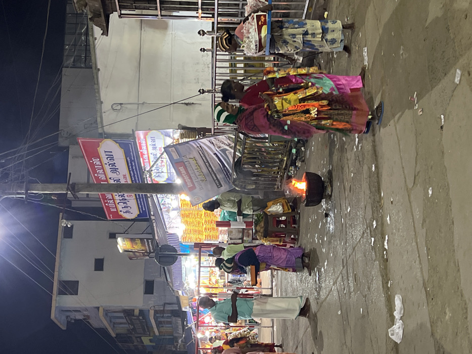 This nighttime image depicts a lively street market scene in Palani, India, characterized by artificial lighting illuminating vendors and shoppers. The ground is paved, appearing somewhat damp and scattered with small debris, reflecting the glow from various light sources.
Several people are visible, mostly women dressed in vibrant traditional Indian sarees, alongside men in dhotis. The activity centers around informal street stalls. A prominent feature in the mid-ground is a black, round pot on the ground with an open, active fire, suggesting on-site cooking or heating of food. Close to this fire, a woman in a pink and multi-colored saree stands near a cart, likely a vendor. Another woman, dressed in a blue and white patterned garment, is seen near a stall displaying a framed picture. Other individuals are present at various stalls, with some goods like coconuts visible.
Overhead, a utility pole dominates the left side of the frame, bearing multiple banners. Visible text from these banners, confirmed by OCR, includes Tamil script such as "மயில்வா விலாஸ்" (Mayilva Villas) and "வாகன விலாஸ்" (Vahana Villas), likely shop names. Advertisements for "திருநெல்லி அல்வா" (Thirunelveli Halwa) and "நெவேலி அல்வா" (Neyveli Halwa) indicate the sale of popular Indian sweets. One banner also carries English text: "Important Information for Irane -PASS MANDATORY FORVENTE," possibly signaling an event or an entry point requiring passes. The overall atmosphere is bustling, typical of an evening market in a South Indian town, especially one known for its religious sites like Palani.