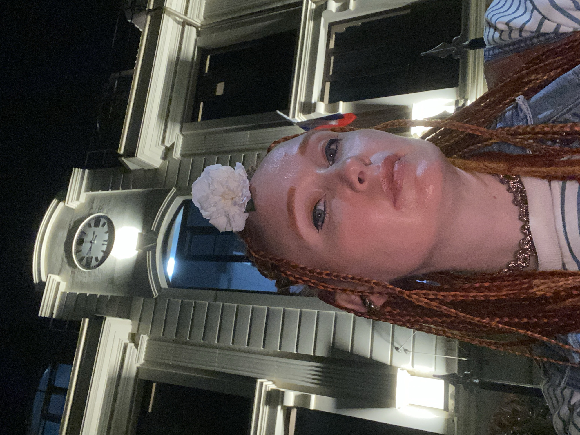 A woman with ginger braids and a white flower on her head is standing at night in front of a building with a clock on it. The building appears to be a historical or government building. The clock shows the time is approximately 11:58. There is a Russian flag visible on the building to the right of the woman. The woman is wearing a denim jacket and a striped shirt. She is looking directly at the camera. The overall scene suggests an urban environment at night.
