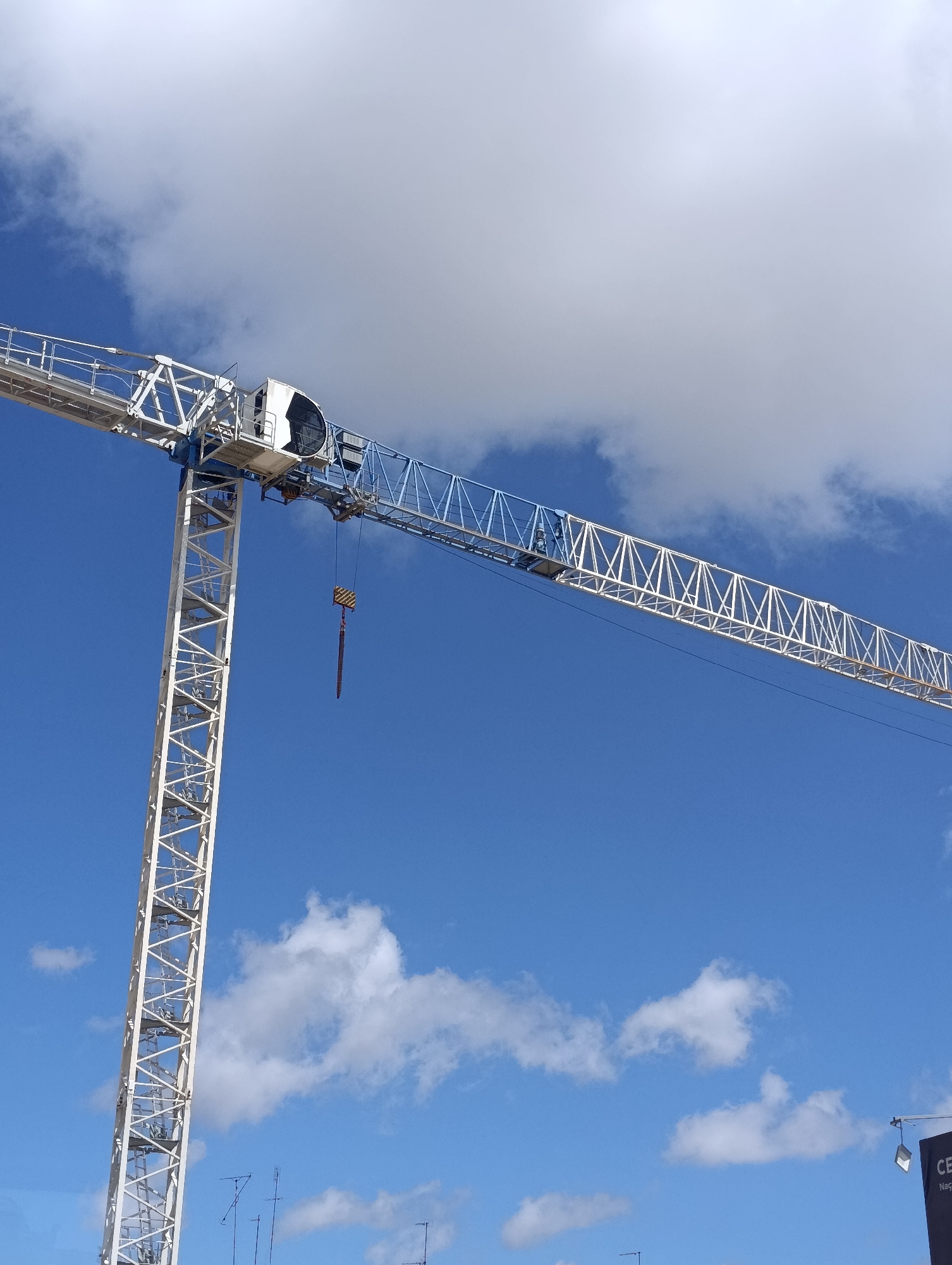 This is a photograph of a large construction crane against a bright blue sky with some white clouds. The crane is predominantly white and blue, with a long horizontal boom extending to the right and a vertical tower to the left. A hook is suspended from the boom. The image appears to be taken from a low angle, looking up at the crane. In the lower right corner, a dark sign with some white lettering is partially visible, possibly indicating a company name or project. The overall scene suggests an active construction site in an urban or suburban setting. While the image itself does not explicitly state the location, Setúbal, Portugal, is a city known for its industrial and construction activities, making it a plausible setting for such a crane.