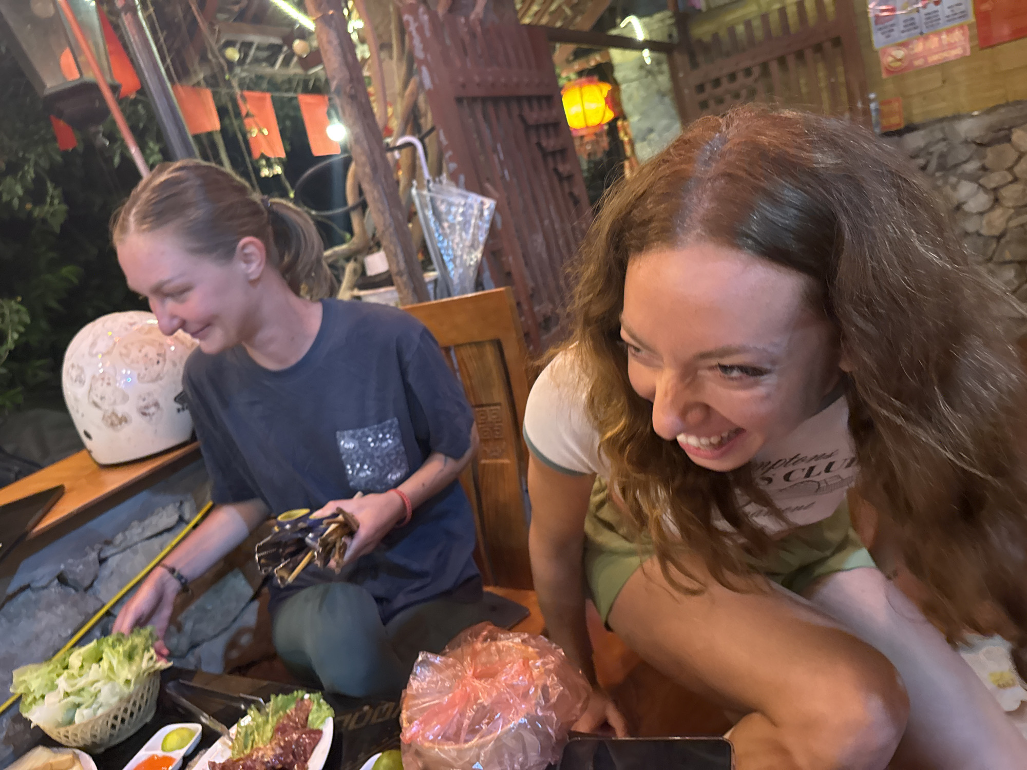 Two women are seated at a table laden with food, likely in the Mèo Vạc Commune of Vietnam, given the context. The woman on the left wears a dark t-shirt with a decorative square and green pants. She has her hair in a ponytail and is handling several dark objects. Next to her, there is a white helmet with a design on it. There are dishes of food, including a basket of lettuce, plates with meat, and small condiment containers. In front of her, there is a covered bowl of food, contained in a pink bag.
The woman on the right is closer to the camera. She has brown, curly hair and is wearing a white shirt with green trim and a green t-shirt. She is smiling broadly. The setting appears to be outdoors or a semi-outdoor space, possibly a restaurant, with trees, lanterns, and other decorative elements visible in the background. The environment is somewhat rustic with wooden elements and possibly stone or brick features.
