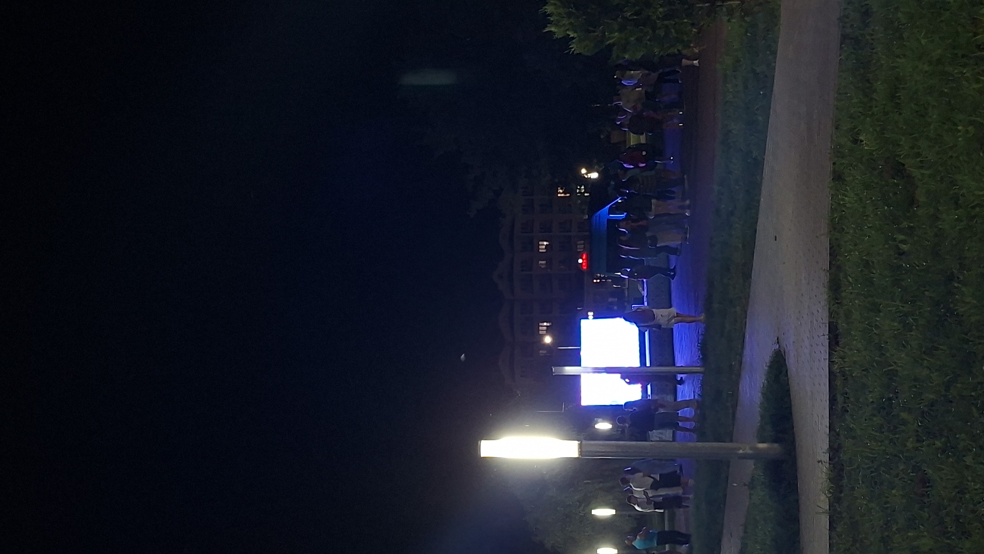 The image depicts a night scene in a public space, likely a park or plaza, in the city of Myadzyel, Belarus. A large, bright LED screen is prominently displayed in the center of the scene, illuminating the surrounding area. A group of people are gathered in front of the screen, some standing and observing, while others appear to be interacting. A light pole with a modern design is visible in the foreground to the left, casting a warm glow. In the background, a multi-story building with illuminated windows is visible, suggesting it is an apartment complex or hotel. The overall atmosphere is one of a public gathering or event, with the screen likely serving as a focal point for entertainment or information. The environment appears to be paved with walkways, and grass is visible in the foreground, indicating a well-maintained public space. The time of day is clearly night due to the darkness of the sky and the artificial lighting.