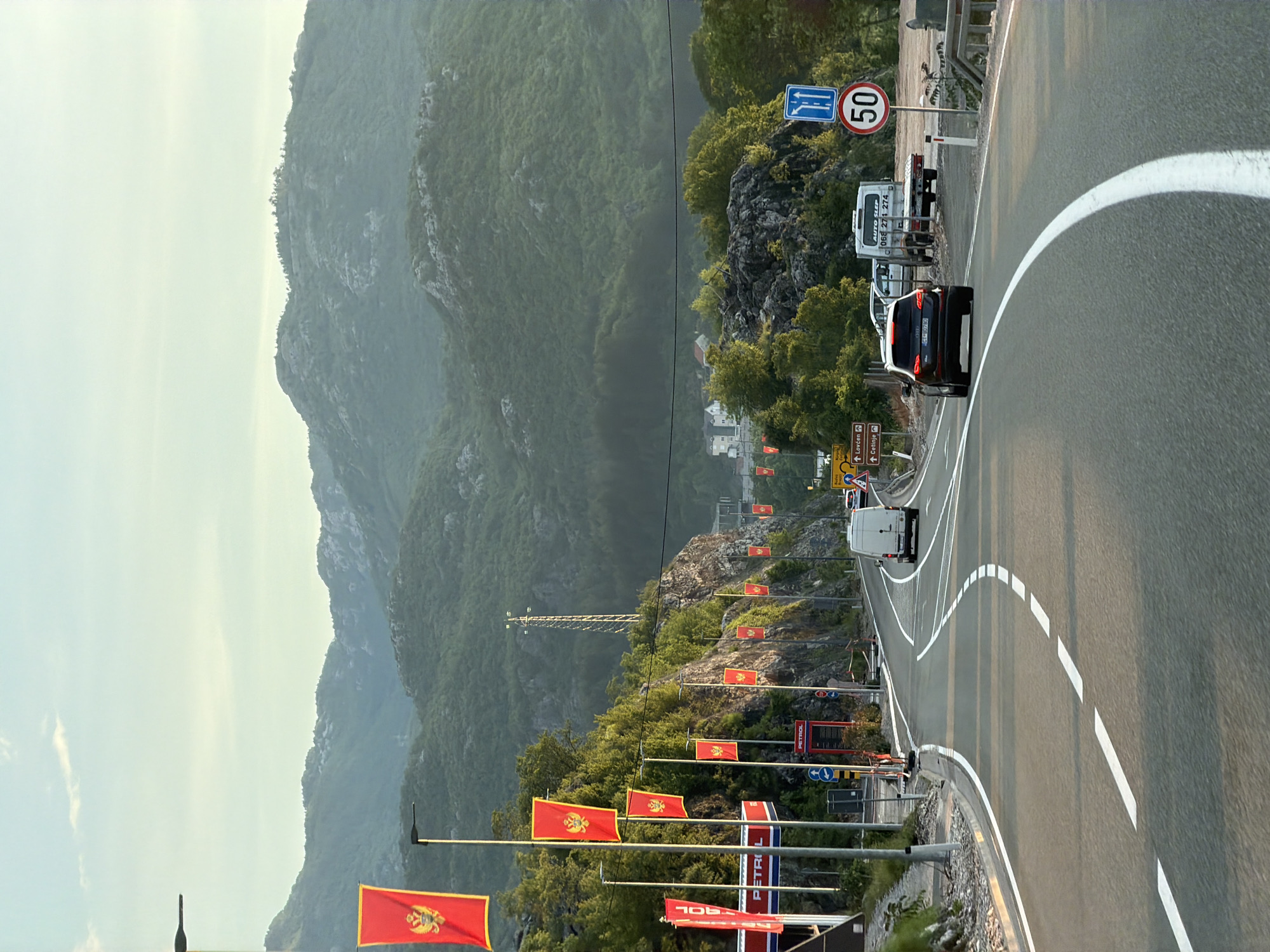 This photograph captures a roadside scene near Crna Greda, Montenegro. A winding asphalt road, marked with solid and dashed white lines, takes center stage, bordered by guardrails and poles adorned with Montenegrin flags. Several vehicles are present on the road, including a dark-colored car and a white van. The signage is also noticeable, including speed limit signs and road signs indicating directions to Lovćen and Cetinje. A petrol station, identified by a "PETROL" sign, is located near the roadside. The backdrop features a mountainous landscape under a slightly hazy sky. A tall electrical tower stands out amidst the greenery.