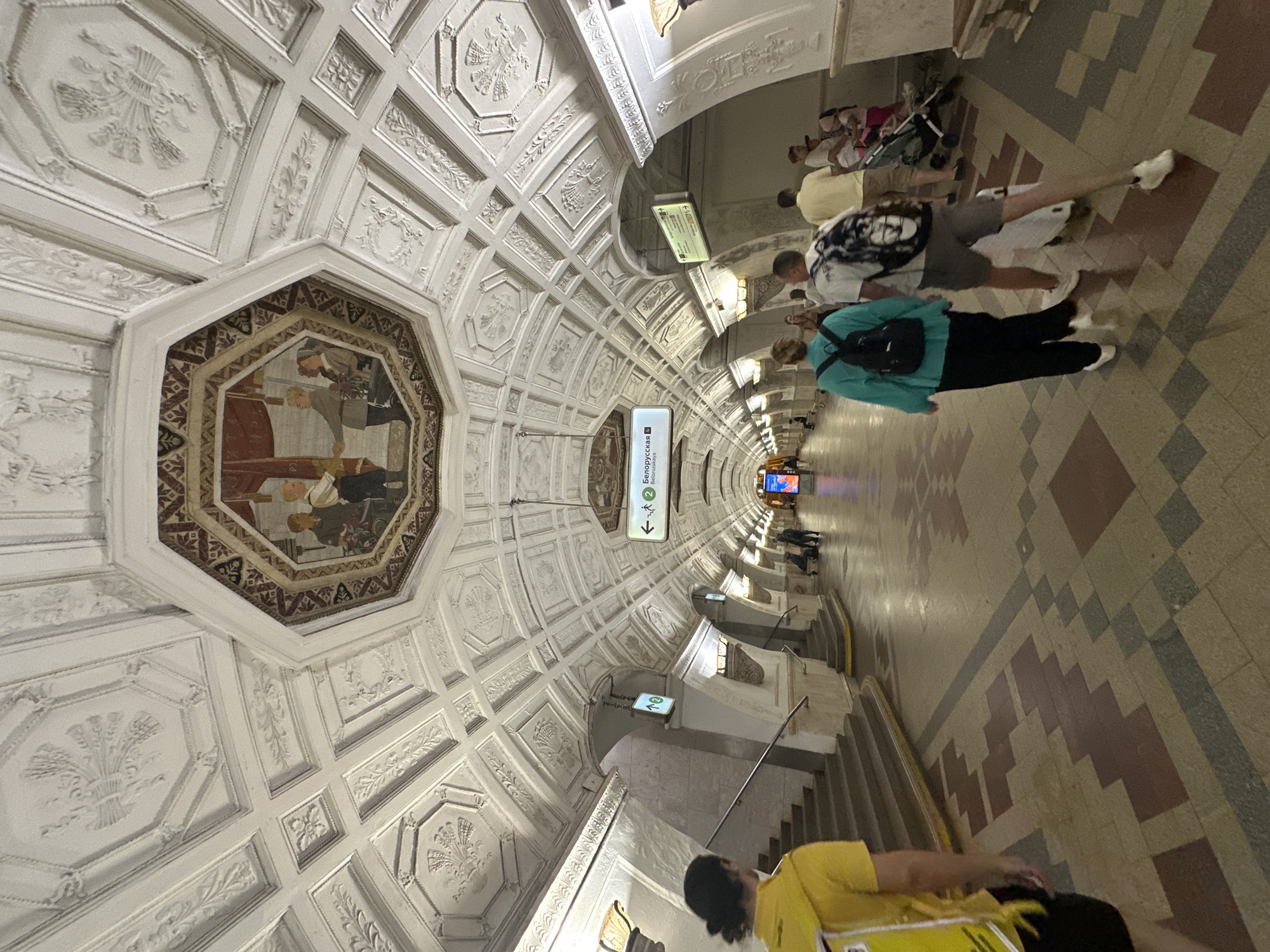 The image shows a wide-angle view of the interior of a Moscow Metro station, likely the Belorusskaya station due to the signage visible. The ceiling is highly ornate, with intricate white stucco moldings and a large octagonal fresco in the center depicting figures in historical attire. The station's architecture features a vaulted ceiling with decorative panels and arched entrances. The floor is paved with geometric patterns in earthy tones. Several people are walking through the station. In the foreground, a person wearing a yellow shirt and black shorts walks away from the camera towards the left. Further down the platform, a group of people are seen walking, including a woman in a teal top and black pants with a backpack, and a man in a white shirt and grey shorts with a backpack. Other individuals and a stroller are also visible further in the distance. The overall scene depicts a busy underground transit environment with a strong emphasis on architectural grandeur and public art.