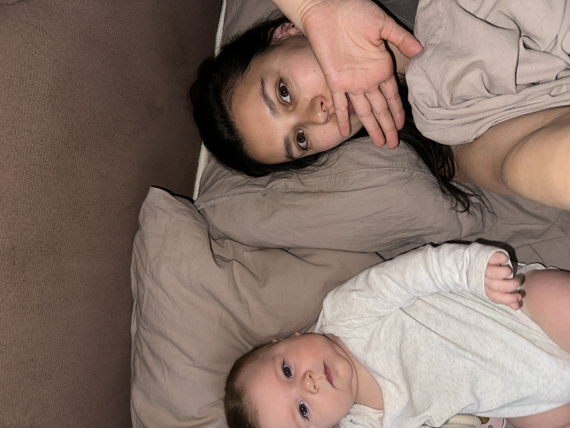 A woman and a baby are lying on a bed. The woman has dark hair and brown eyes and is lying on her back with her hand near her face. The baby is also lying on its back, wearing a light-colored shirt. The bed has taupe-colored sheets and pillows. The location is likely indoors, with a brown couch or wall visible in the background. The image's origin or location within Chaikovsky, Russia, cannot be determined from the provided information.