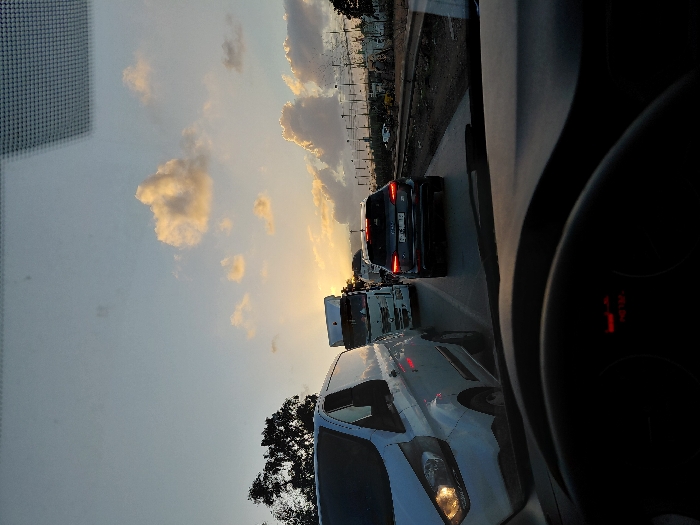 A view from inside a car stuck in traffic at sunset. Several vehicles are visible in the adjacent lane, including a white van, a truck, and a dark-colored car. The sky is filled with clouds, and the sun is positioned near the horizon, creating a warm glow. The dashboard and steering wheel are partially visible in the foreground, with some indicator lights illuminated.